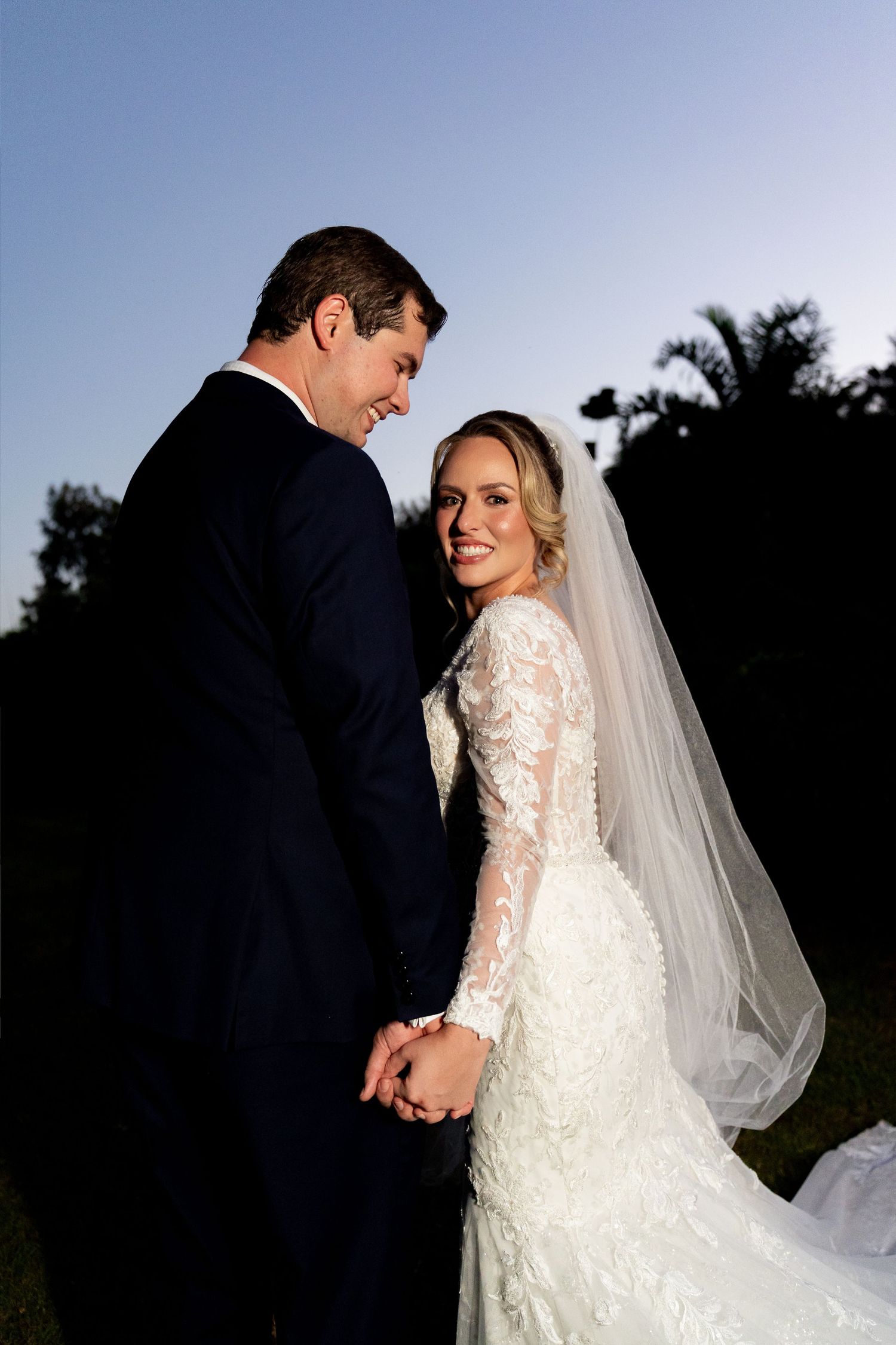 Bride in lace long-sleeve wedding gown and veil smiles over shoulder while holding hands with groom in navy suit outdoors.