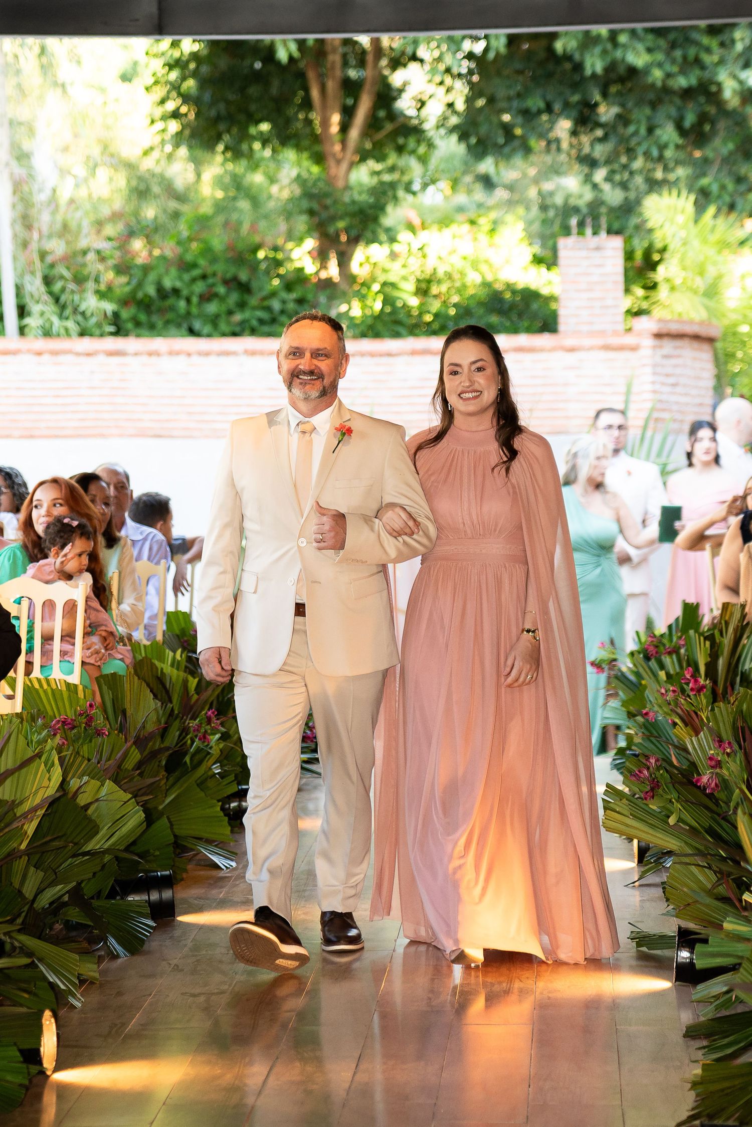 A couple walks down a wedding aisle, man in beige suit, woman in pink gown, surrounded by tropical floral arrangements.
