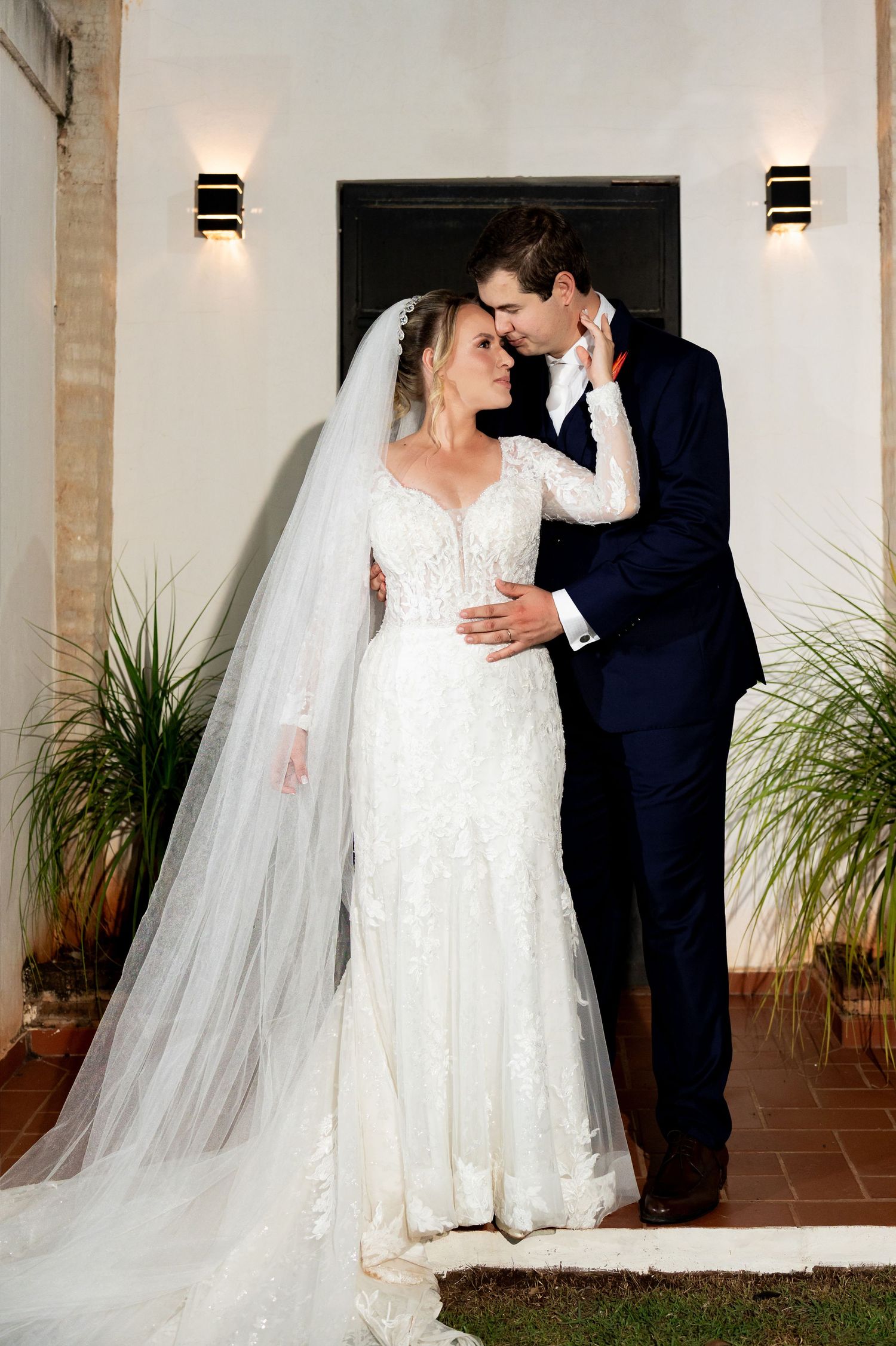 Bride in lace gown and veil embraces groom in navy suit outside elegant venue with wall sconces.