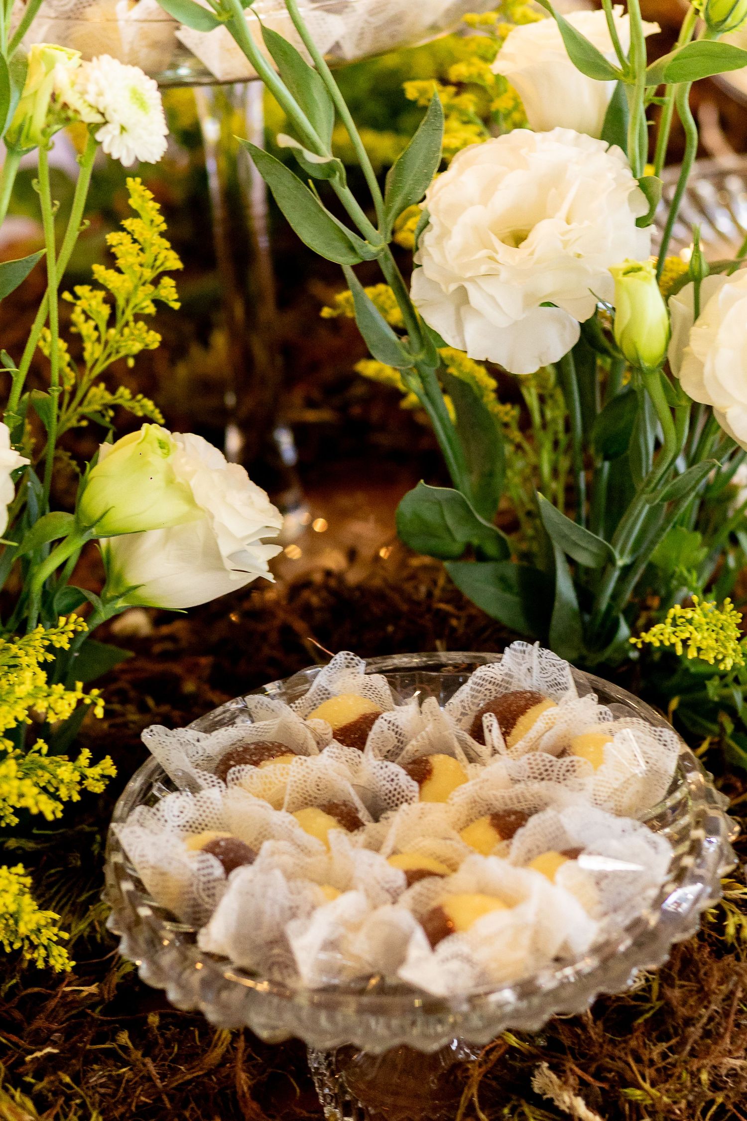Crystal dish of wrapped candies displayed among white lisianthus and yellow mimosa flowers at an elegant event.