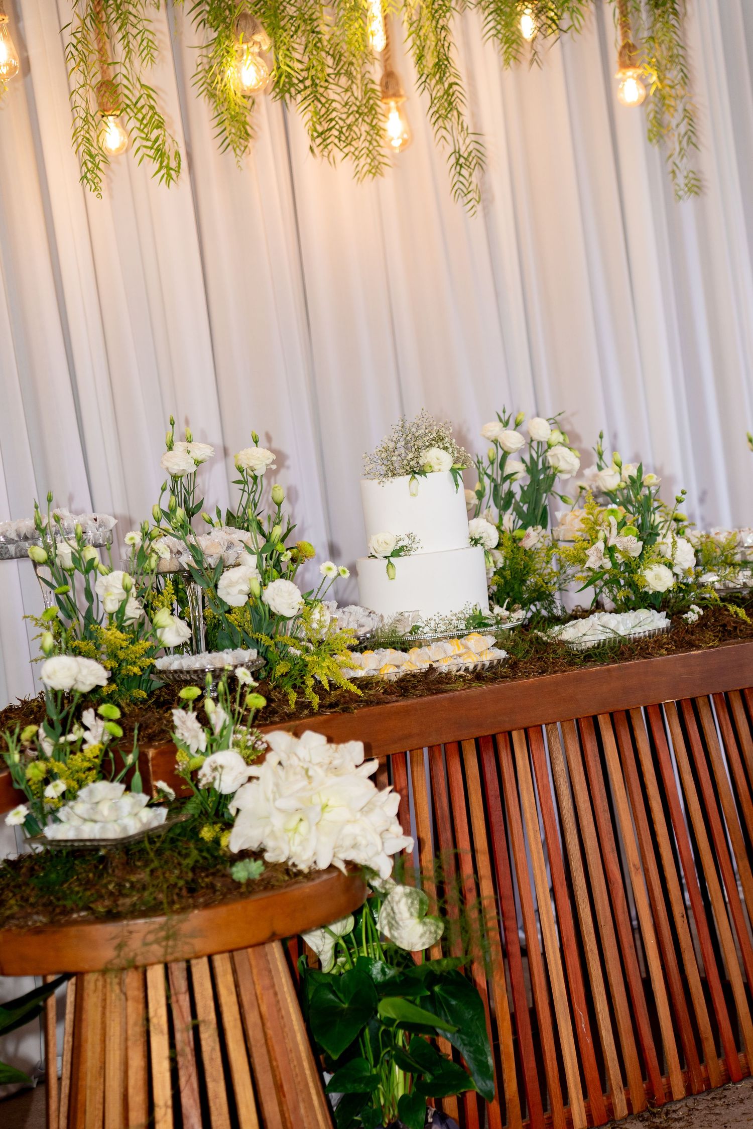 Elegant wedding cake table with white flowers, greenery, and warm Edison bulb lighting against a white draped backdrop.