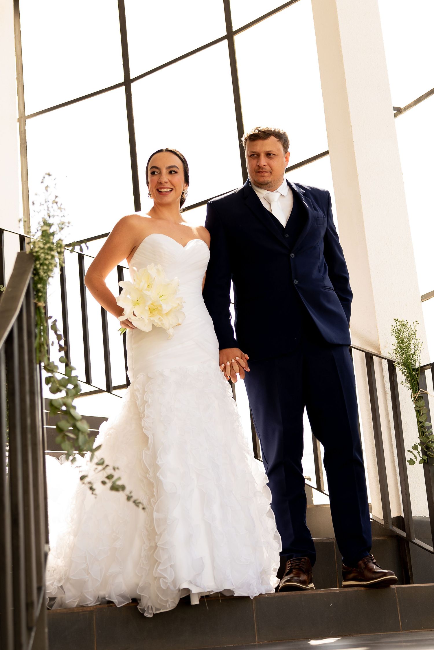 Bride in white mermaid gown holding bouquet stands with groom in navy suit on staircase at wedding venue.