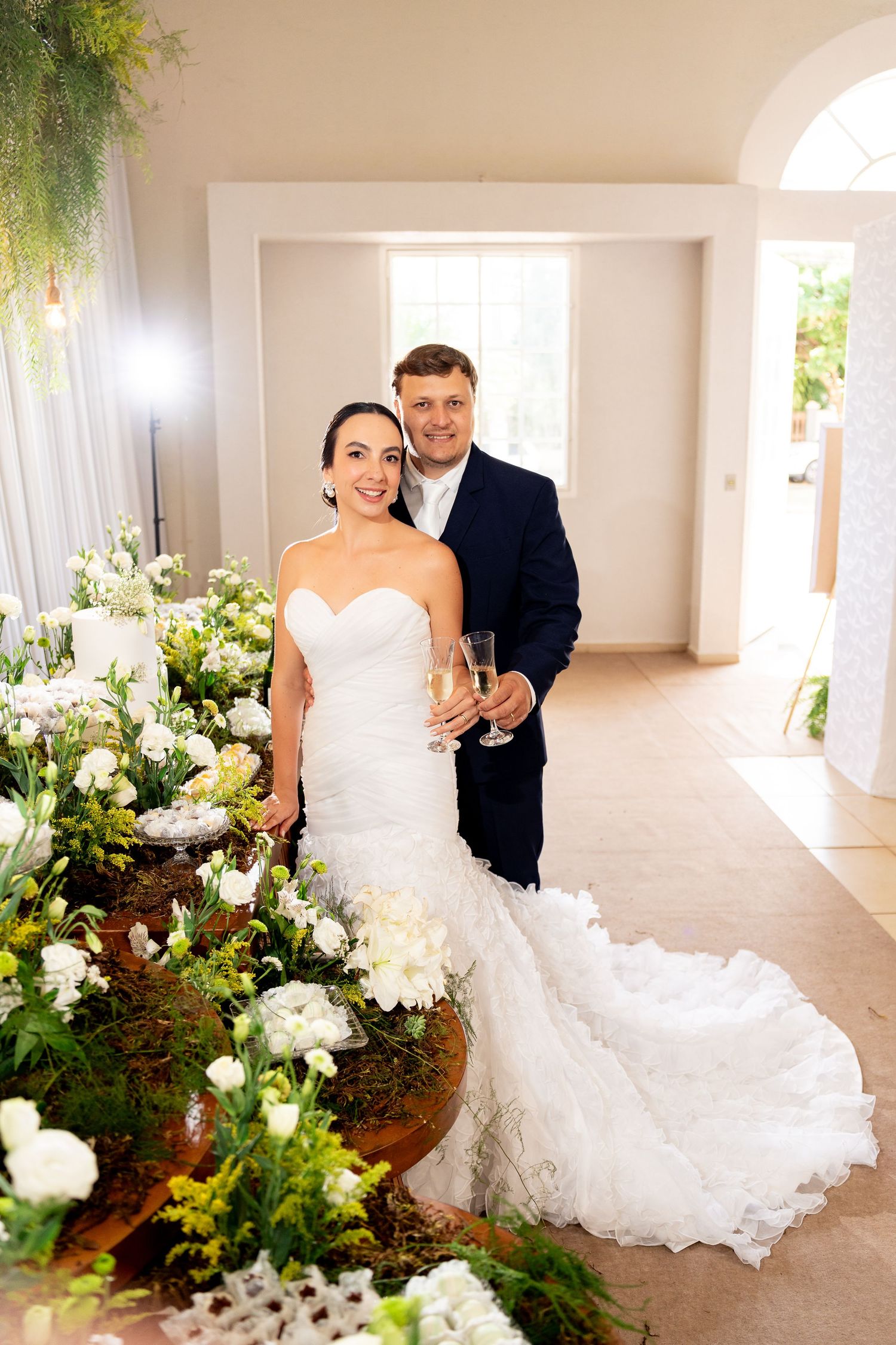 Bride in white mermaid gown and groom in navy suit pose with champagne beside lush floral arrangements at elegant wedding venue.