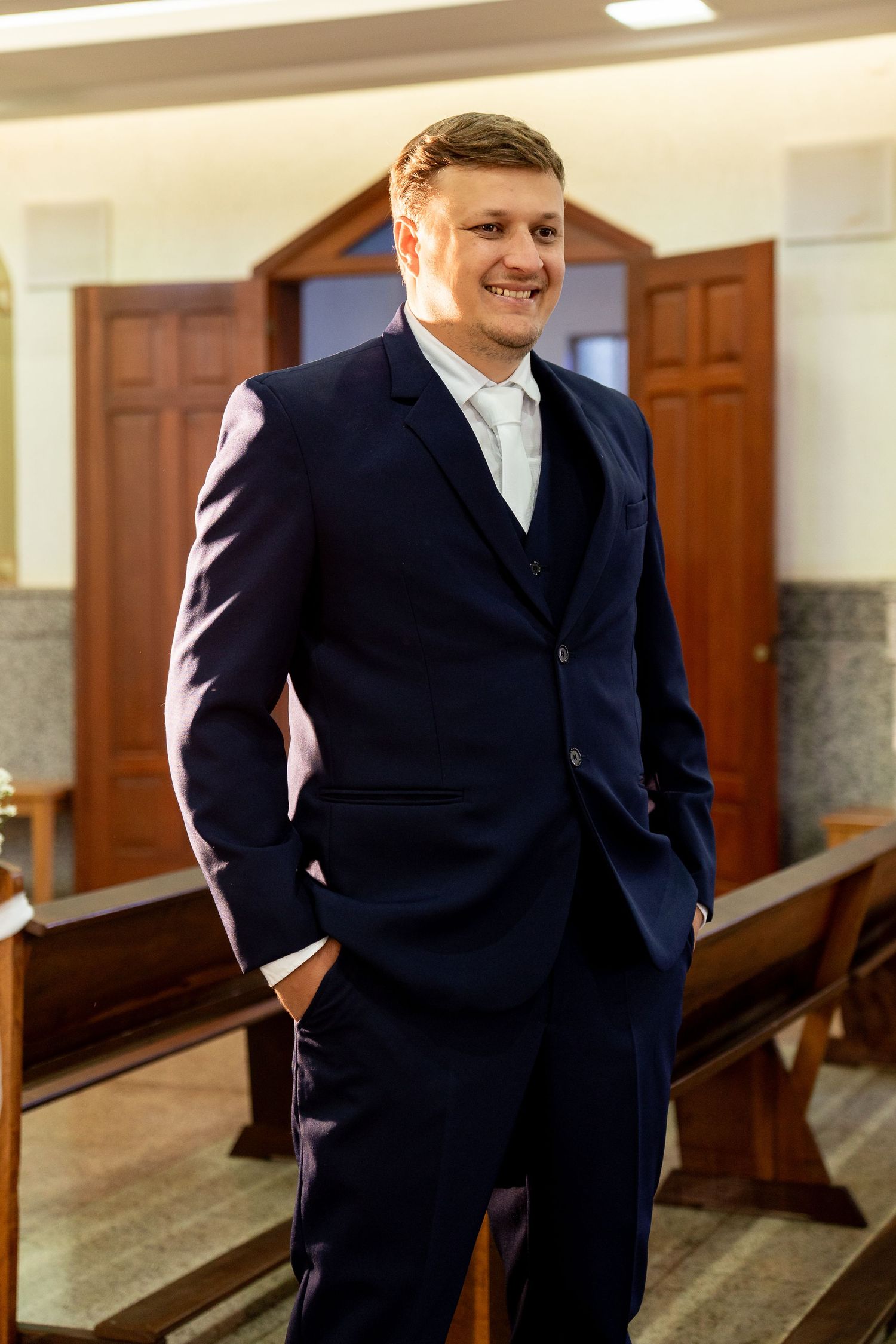 Smiling groom in navy blue three-piece suit standing in a church with wooden pews and doors in background.