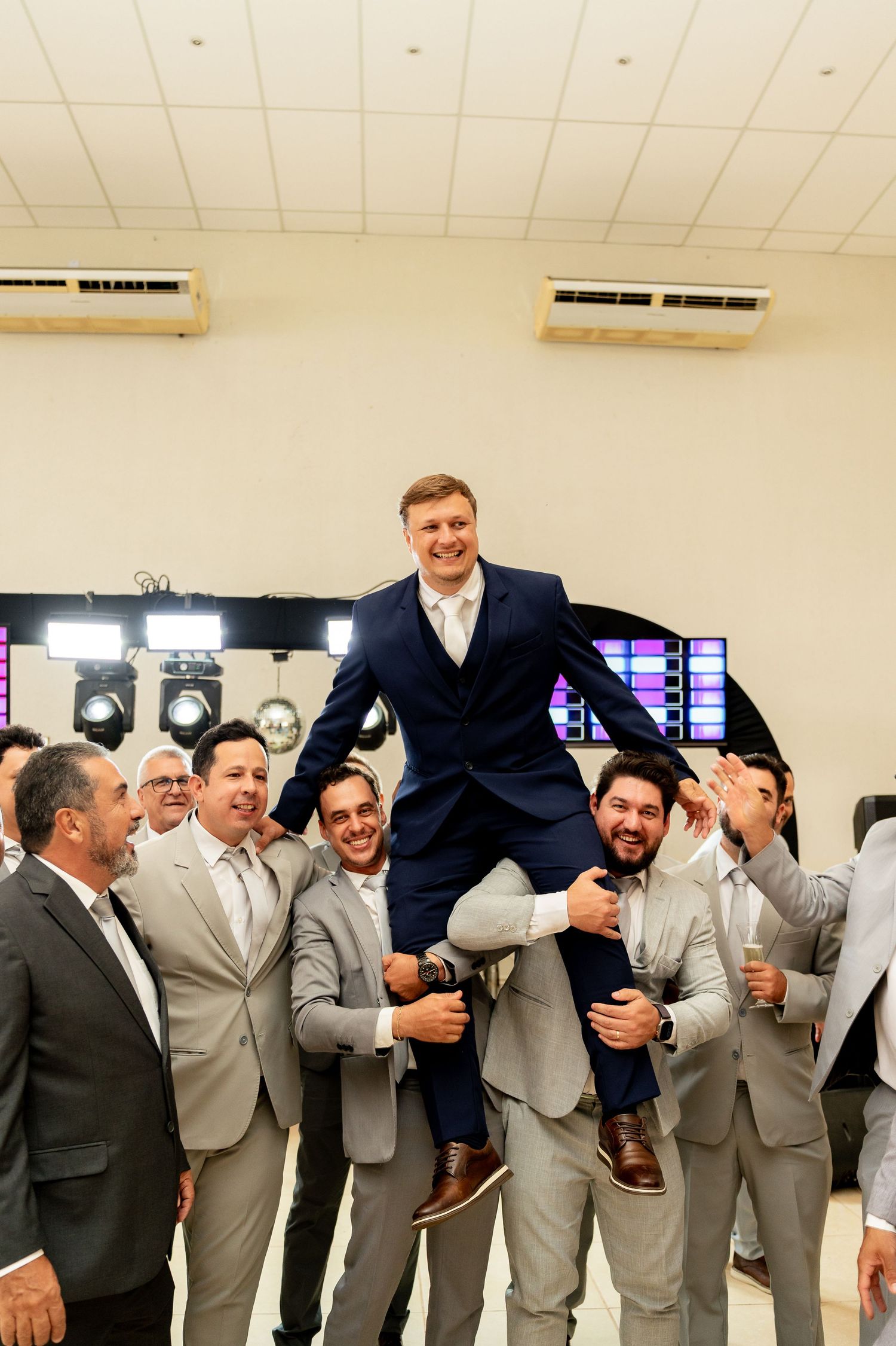 Groom in navy suit lifted by groomsmen in gray suits during joyful wedding celebration indoors.
