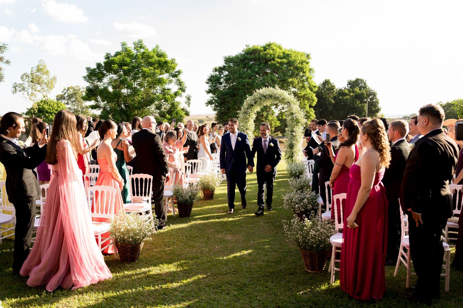 Couple walks down outdoor wedding aisle under floral arch, guests in pink and black attire applaud on lush green lawn.