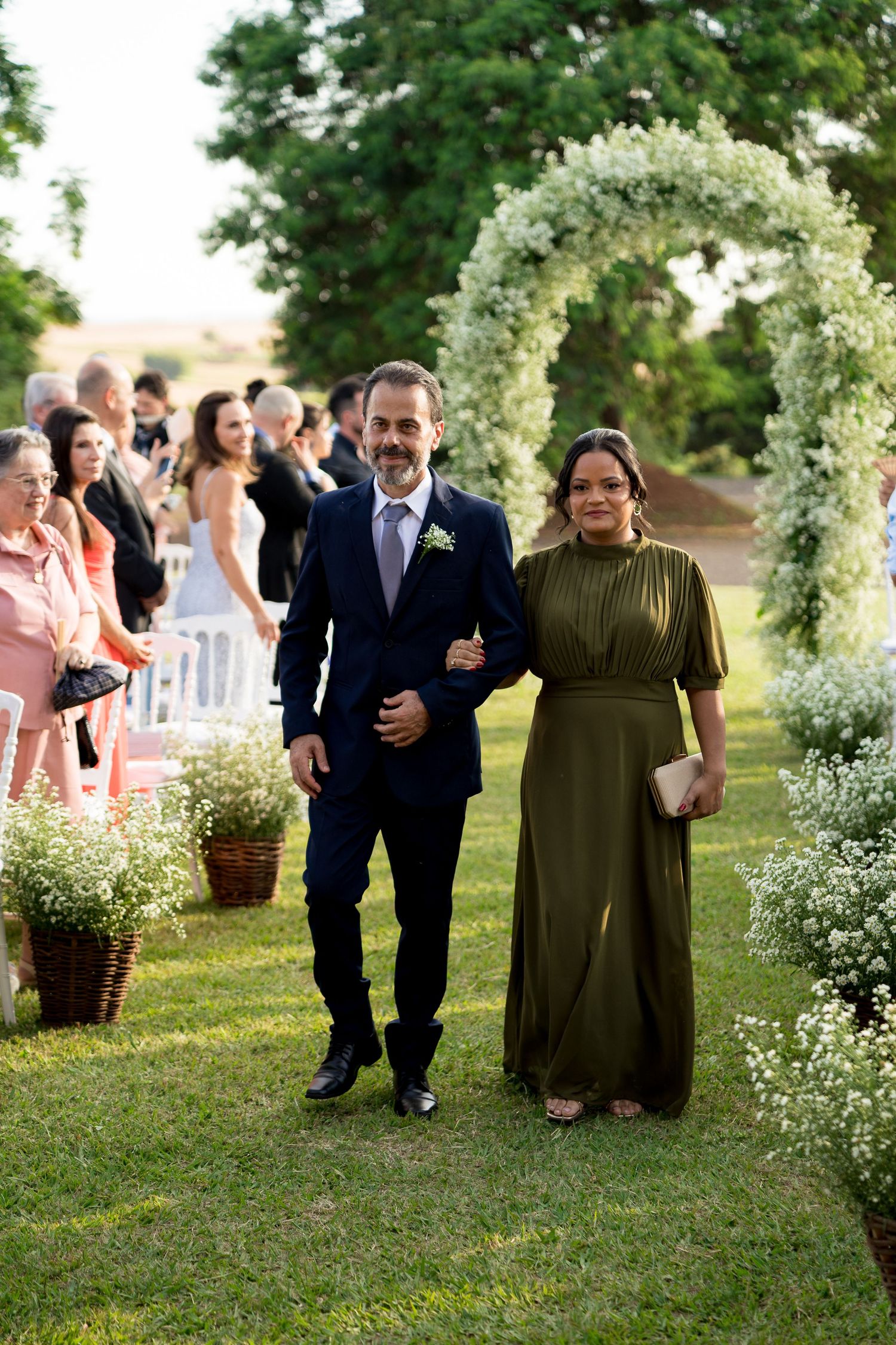 Couple walks down floral arch aisle at outdoor wedding, man in navy suit, woman in olive green gown.