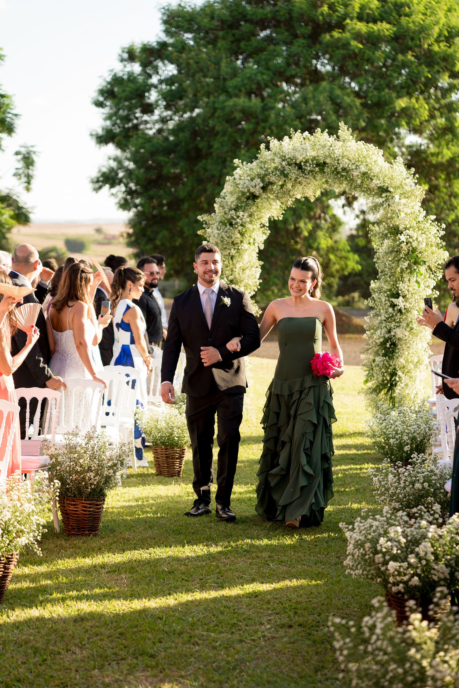 A couple walks down a floral arch-adorned outdoor wedding aisle, guests seated on either side in a lush garden setting.