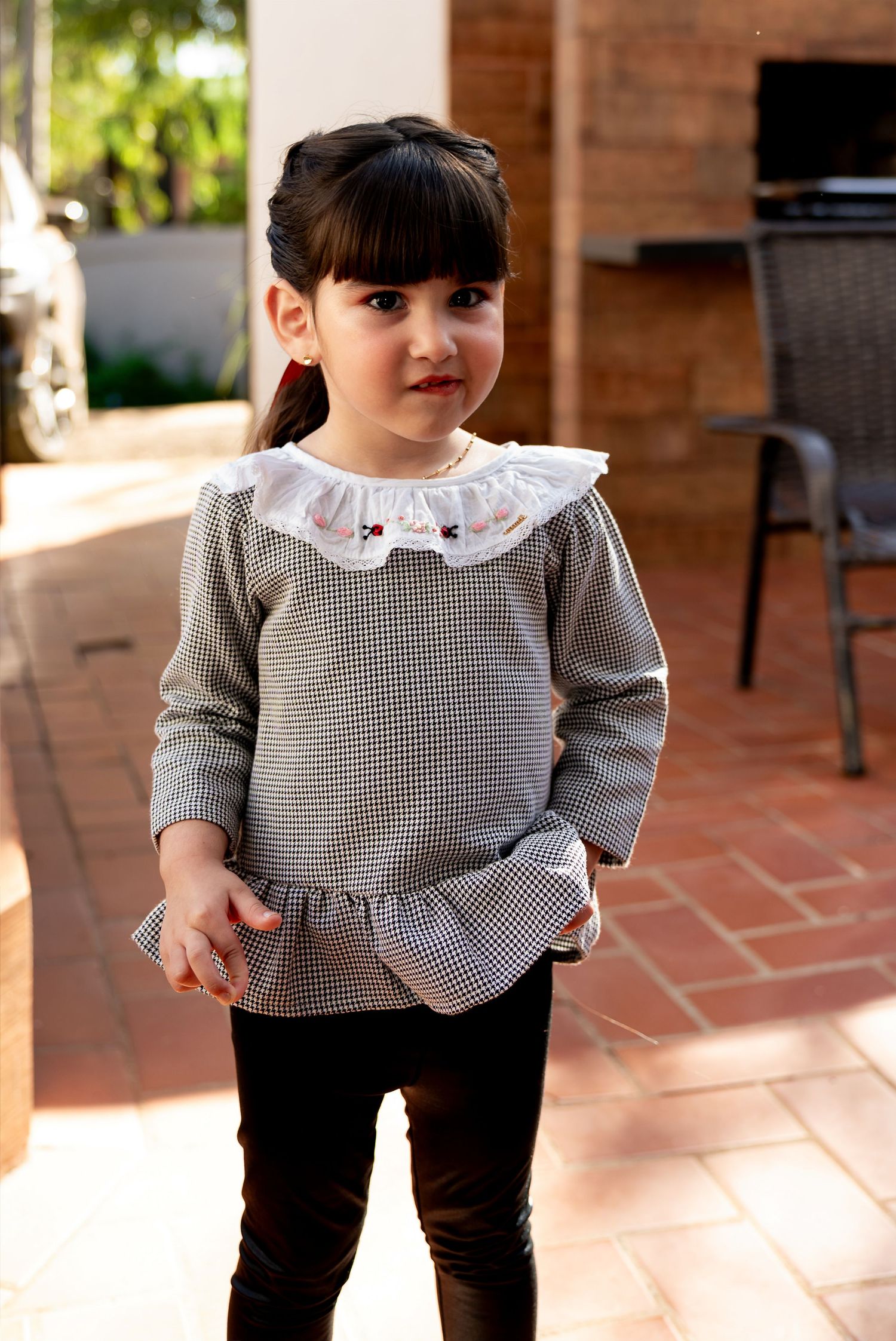 Young girl in checkered top with white ruffle collar and black leggings standing on a brick patio.