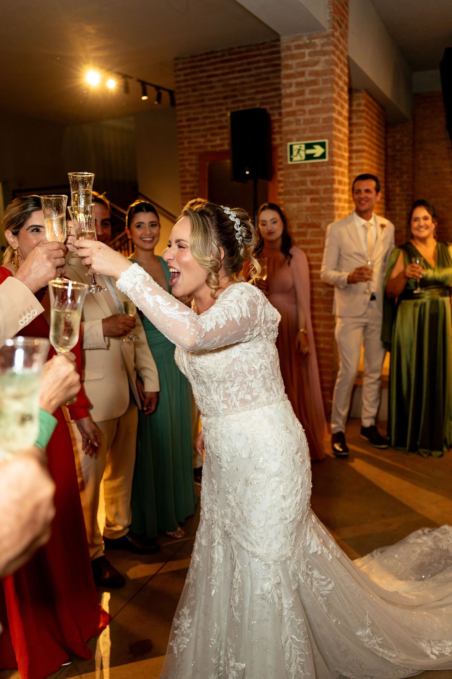 Bride in white lace gown toasting champagne with guests at wedding reception in rustic brick venue.