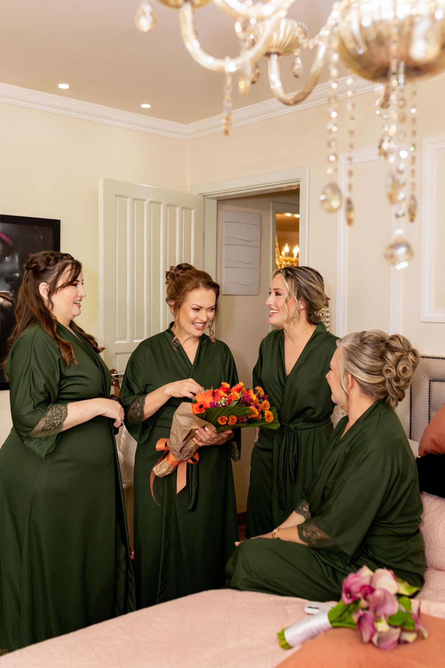 Bridesmaids in green robes gather in elegant room, admiring colorful bridal bouquet before wedding ceremony.