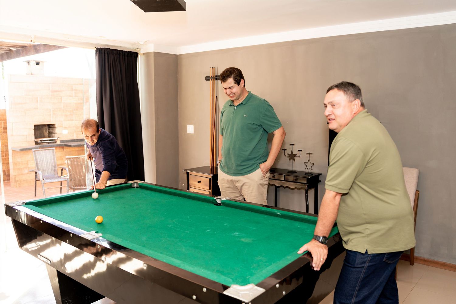 Three men playing pool on a green billiard table in a modern game room with large windows.
