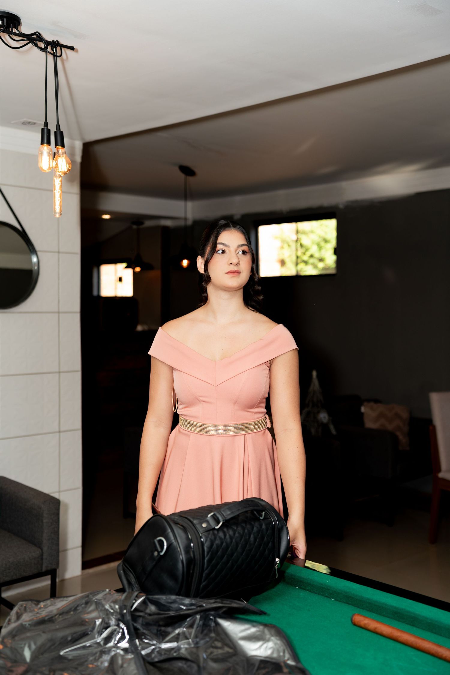 Young woman in pink off-shoulder dress stands near a pool table with a black handbag in a modern interior.