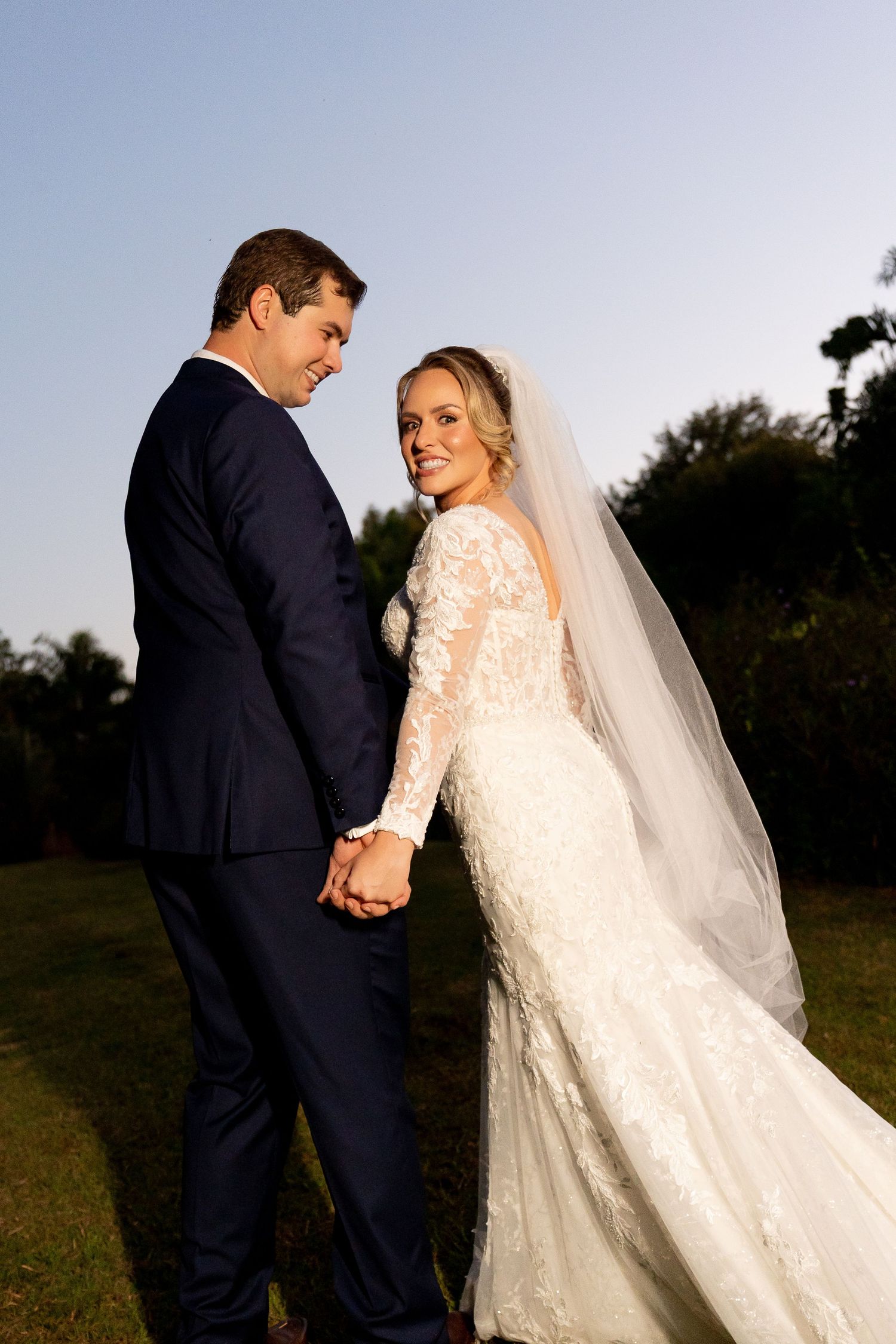 Bride in lace gown with veil holds hands with groom in navy suit, smiling over shoulder outdoors at golden hour.