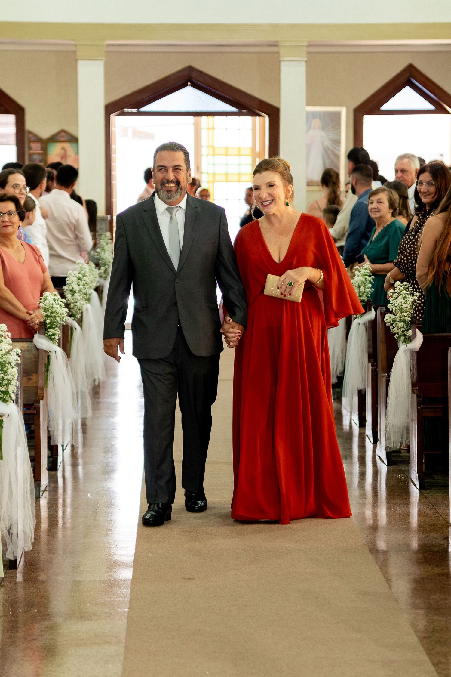 Couple walking down church aisle, woman in red gown and man in gray suit, smiling at wedding ceremony.