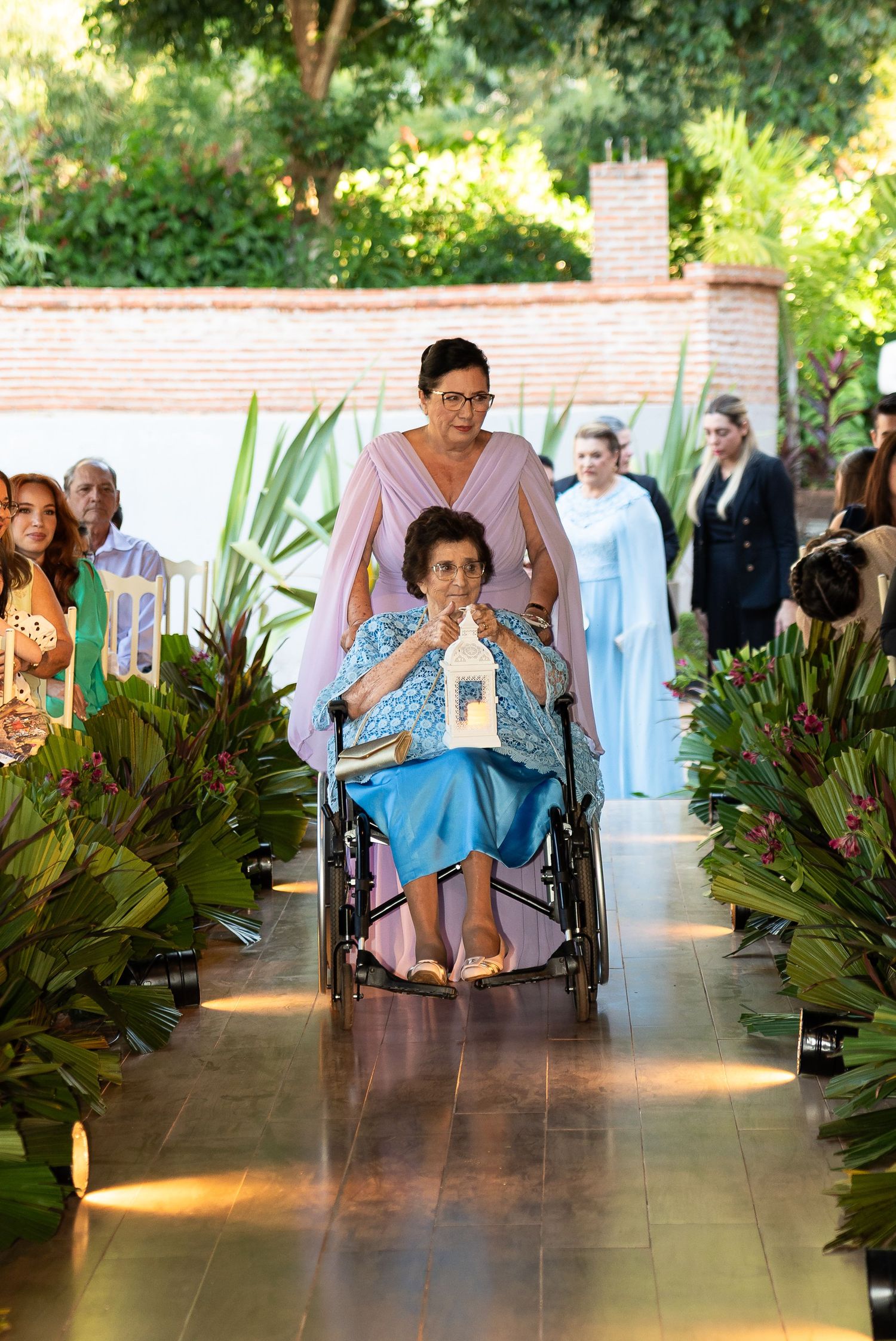 Elderly woman in wheelchair being pushed down floral aisle at outdoor wedding ceremony by woman in pink dress.