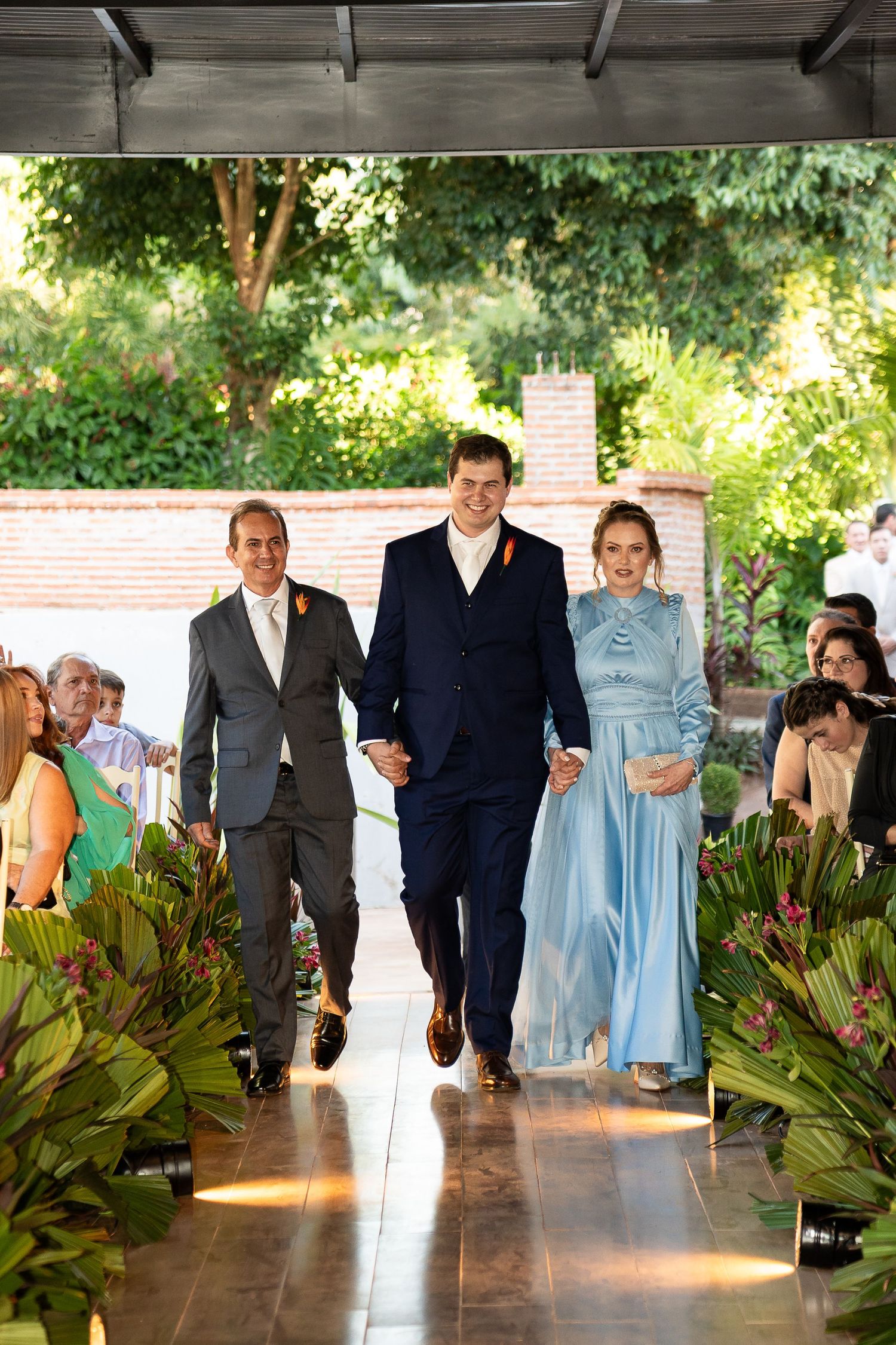 Three elegantly dressed wedding guests walk down a floral-lined aisle at an outdoor garden ceremony.