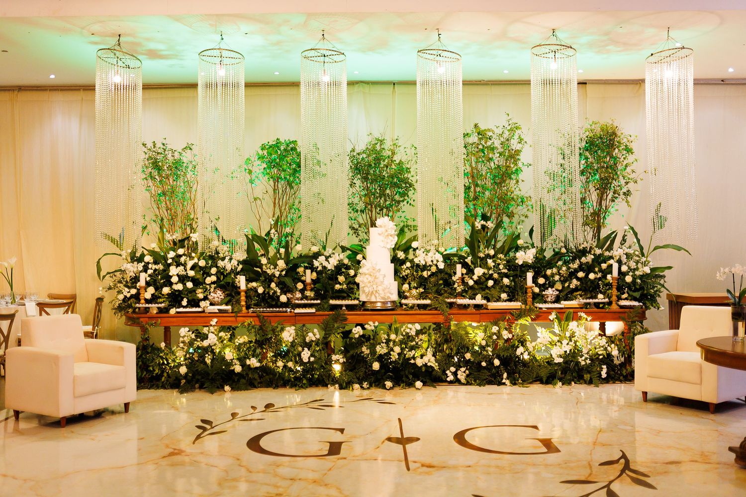 Elegant wedding reception hall with lush floral backdrop, white cake, hanging crystals, and monogram G+G on marble floor.