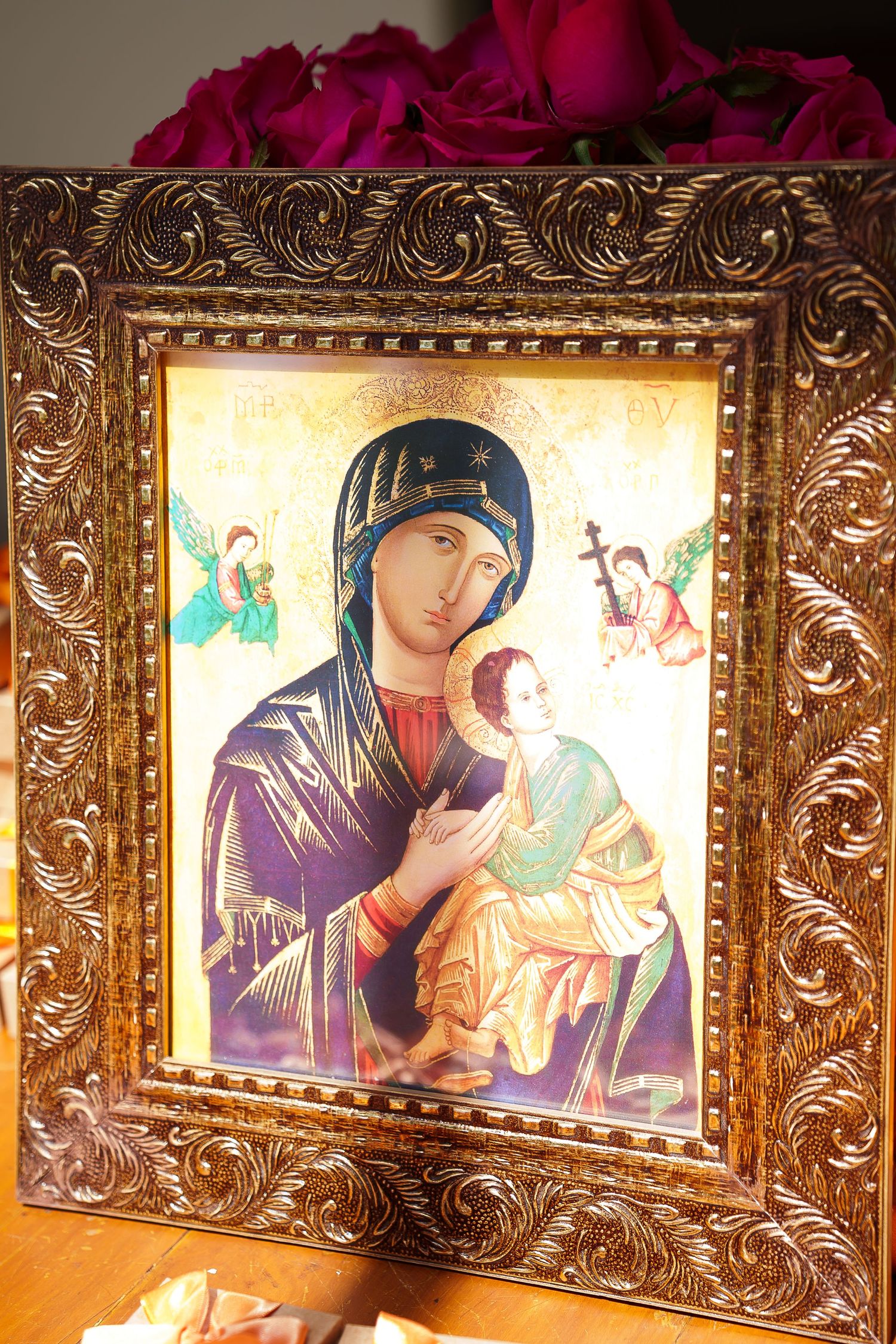 Ornate gold-framed Orthodox icon of Virgin Mary holding baby Jesus with angels, displayed on wooden surface with roses.