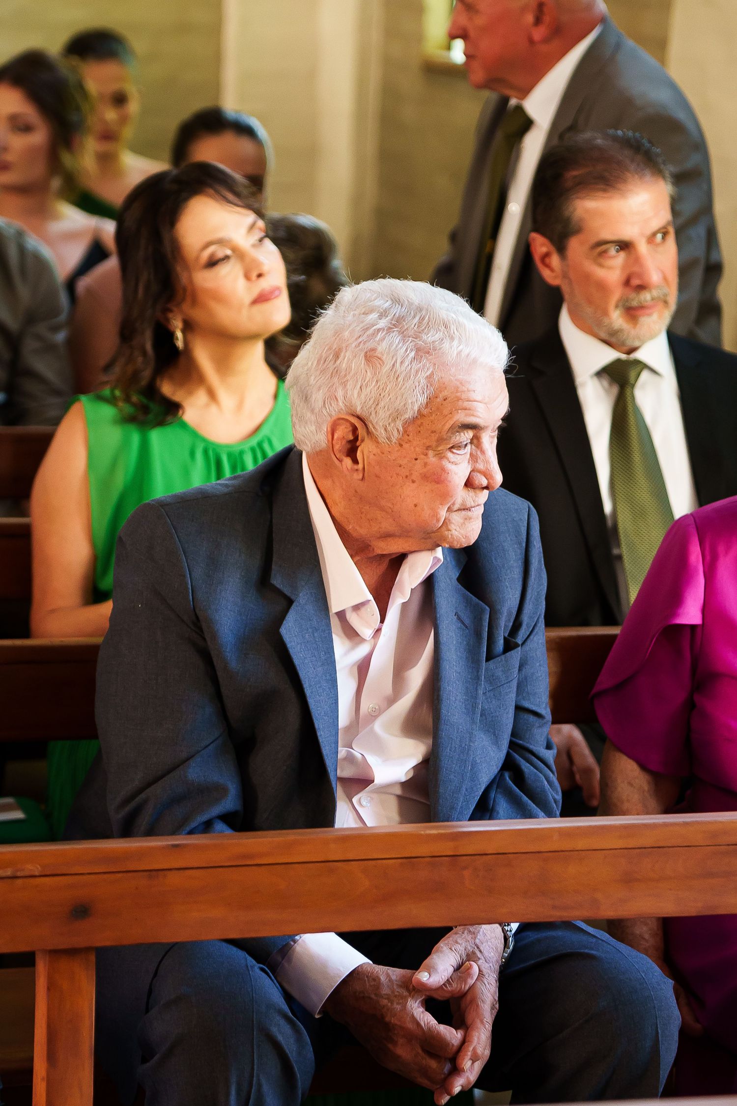 Elderly man in blue suit sits in church pew during a formal ceremony with other guests seated behind him.