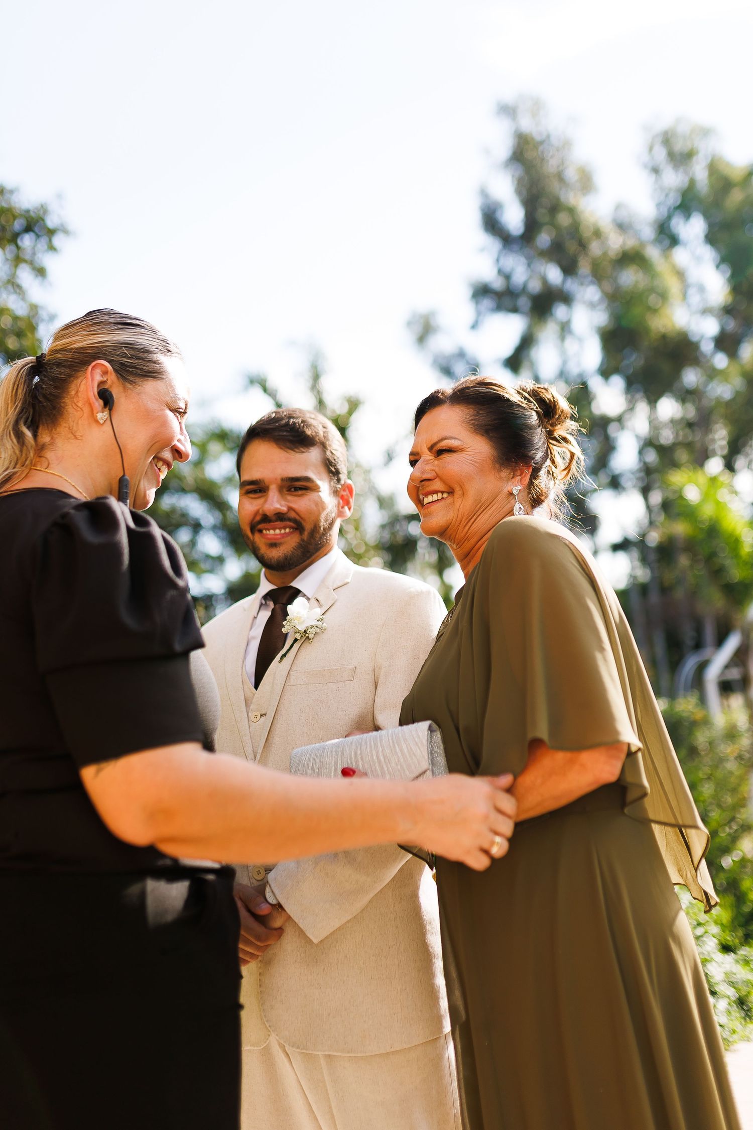 Outdoor wedding ceremony with groom in beige suit, officiant in black, and guest in olive dress laughing together.