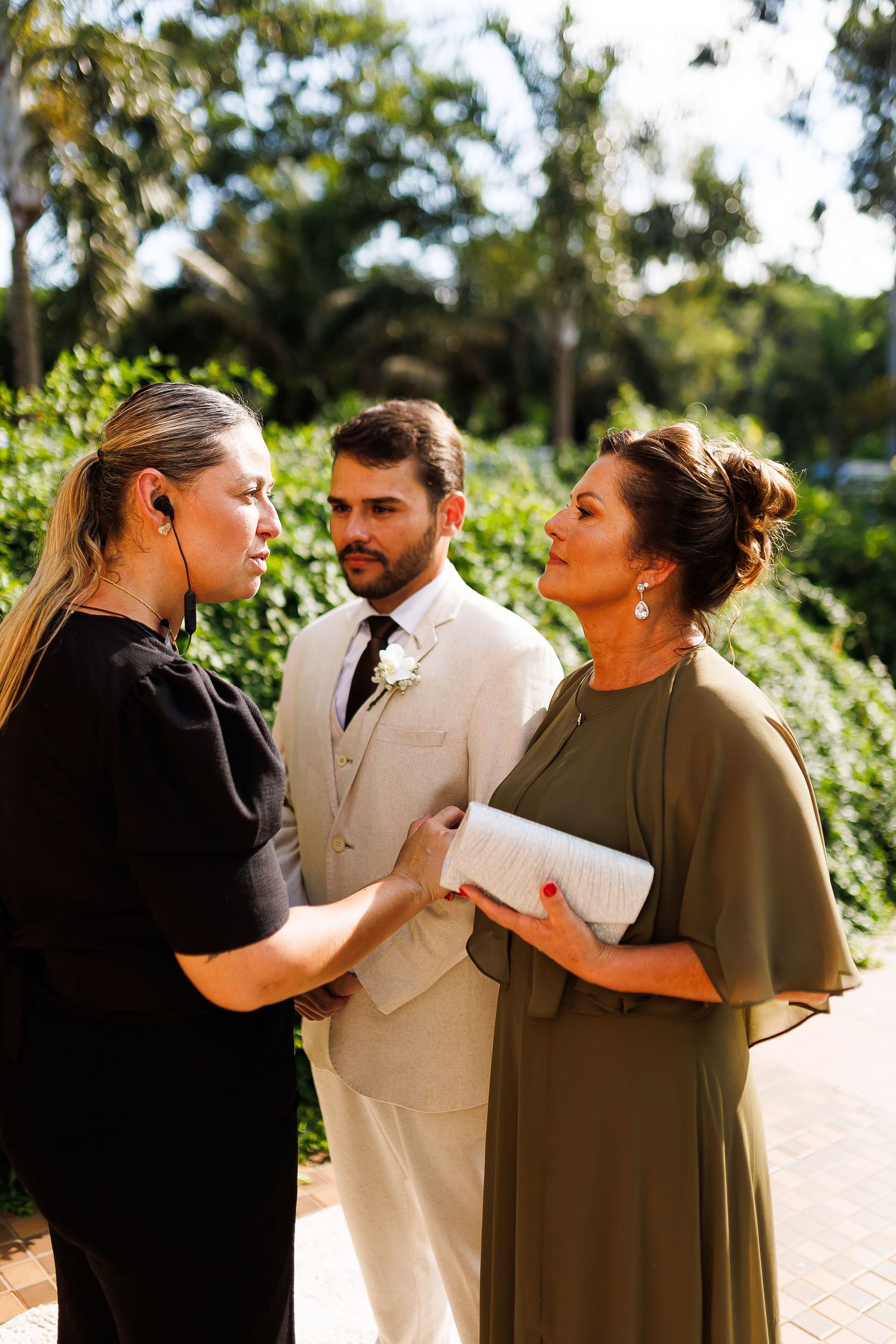 A groom in a beige suit stands between two women during an outdoor wedding ceremony surrounded by lush greenery.