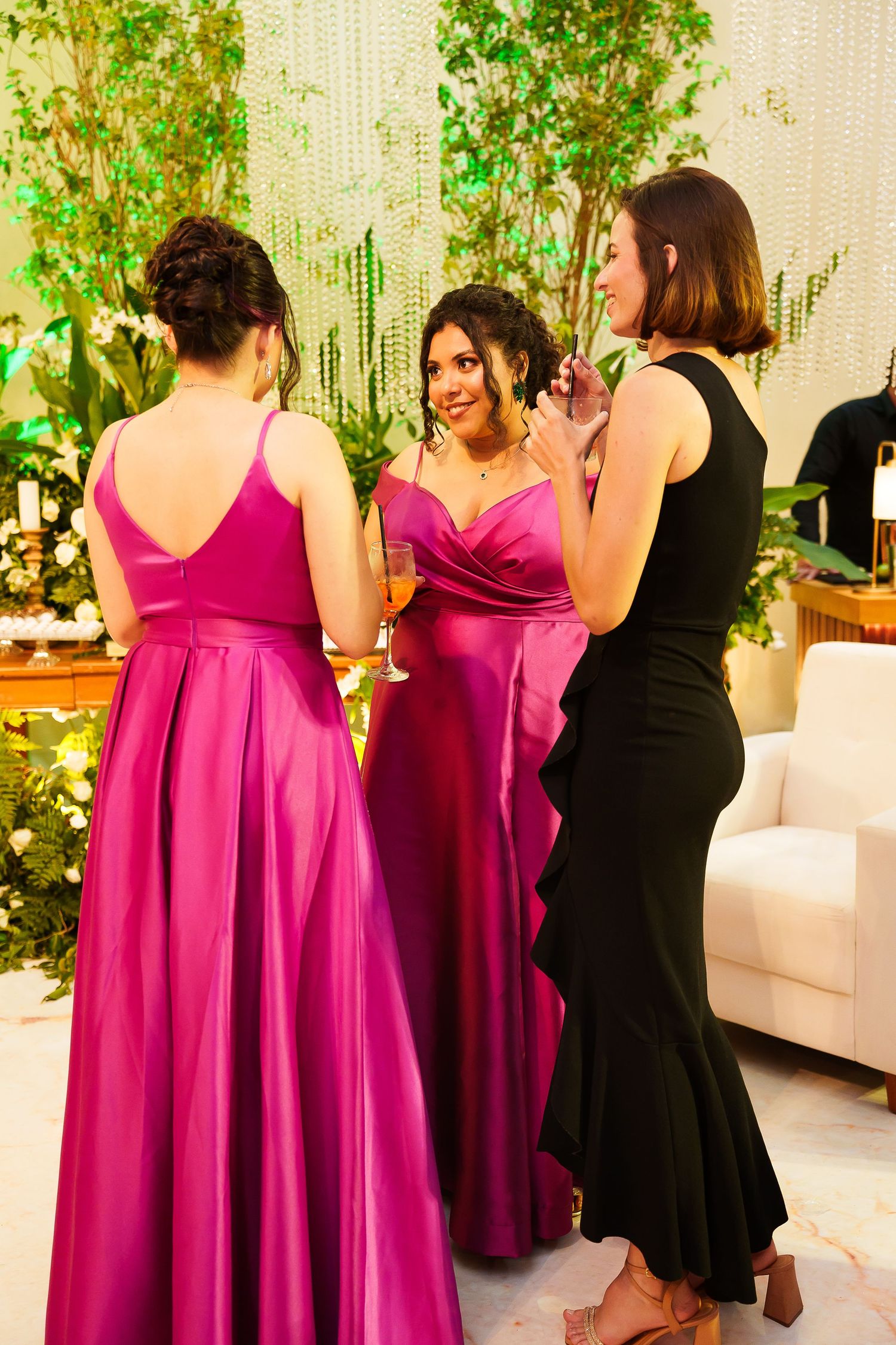 Three women in formal attire, two in magenta gowns and one in black dress, socializing at an elegant event.