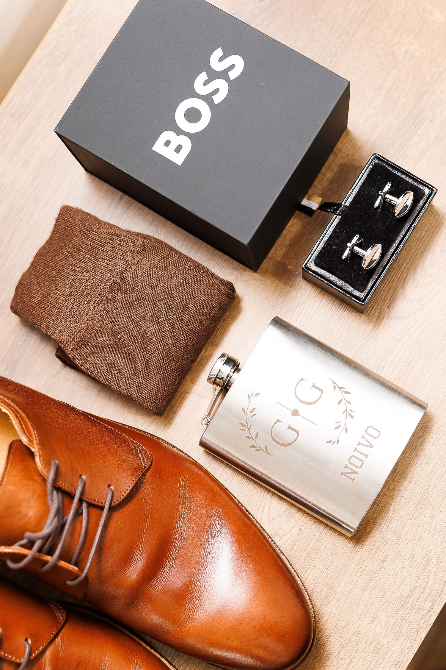 Brown leather dress shoes, BOSS socks, cufflinks, and an engraved silver flask arranged on a wooden surface.