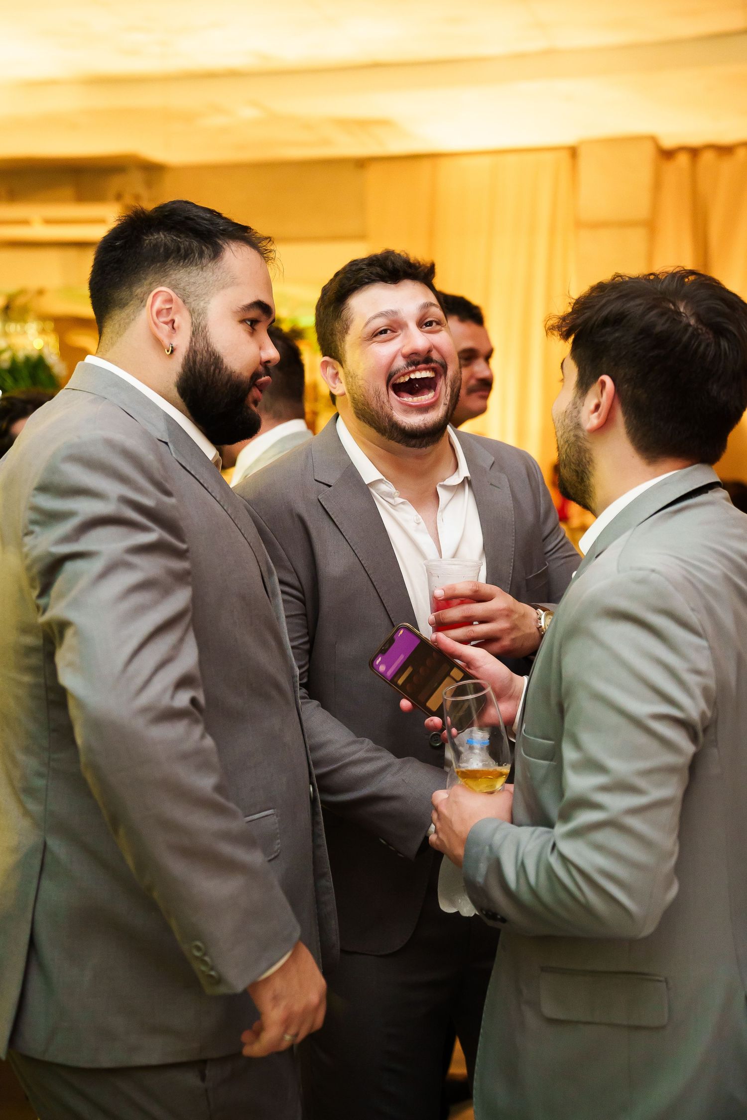 Three men in gray suits laughing and socializing at a formal event, holding drinks.