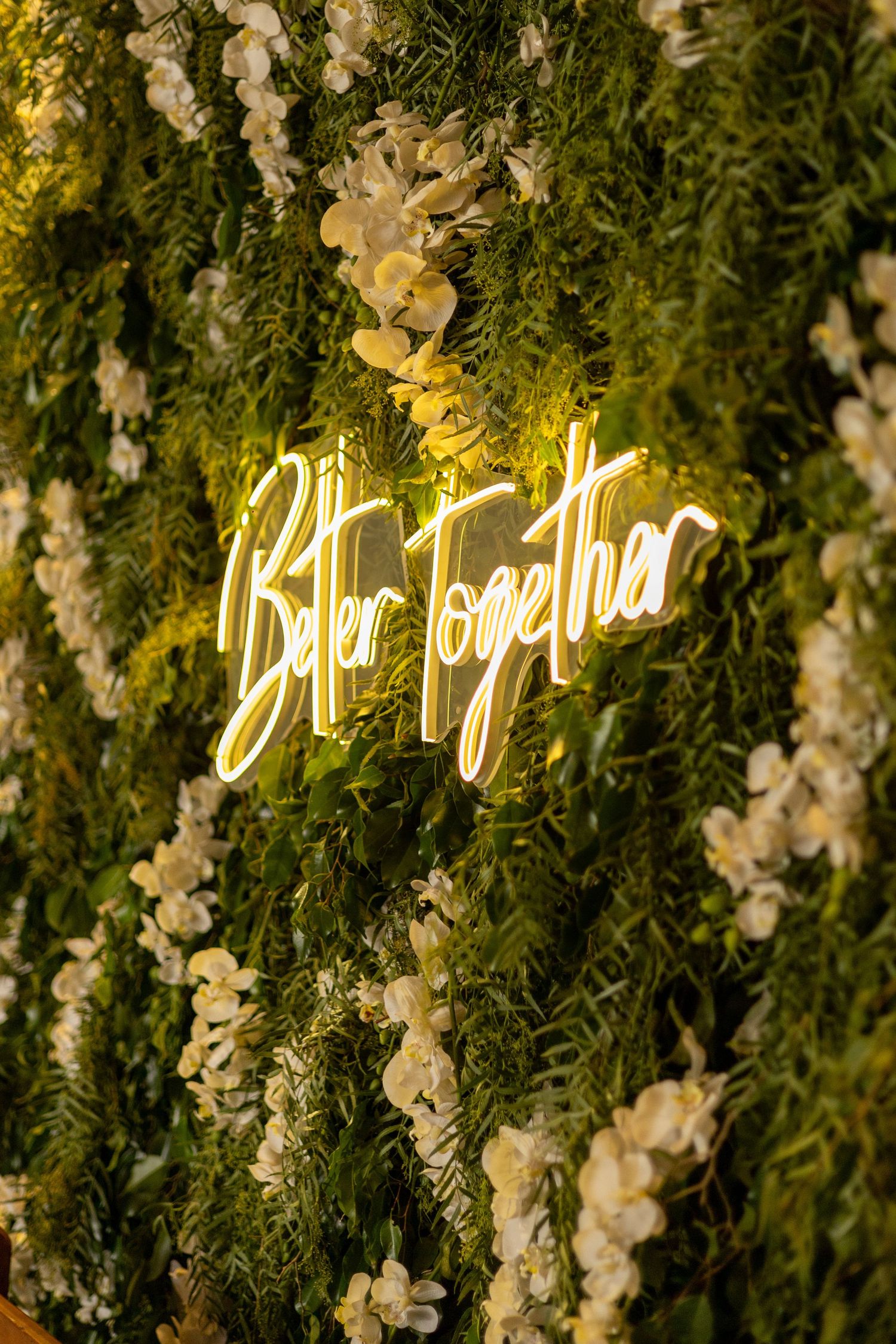 Glowing yellow neon sign reading Better Together on a lush green floral wall with white flowers.
