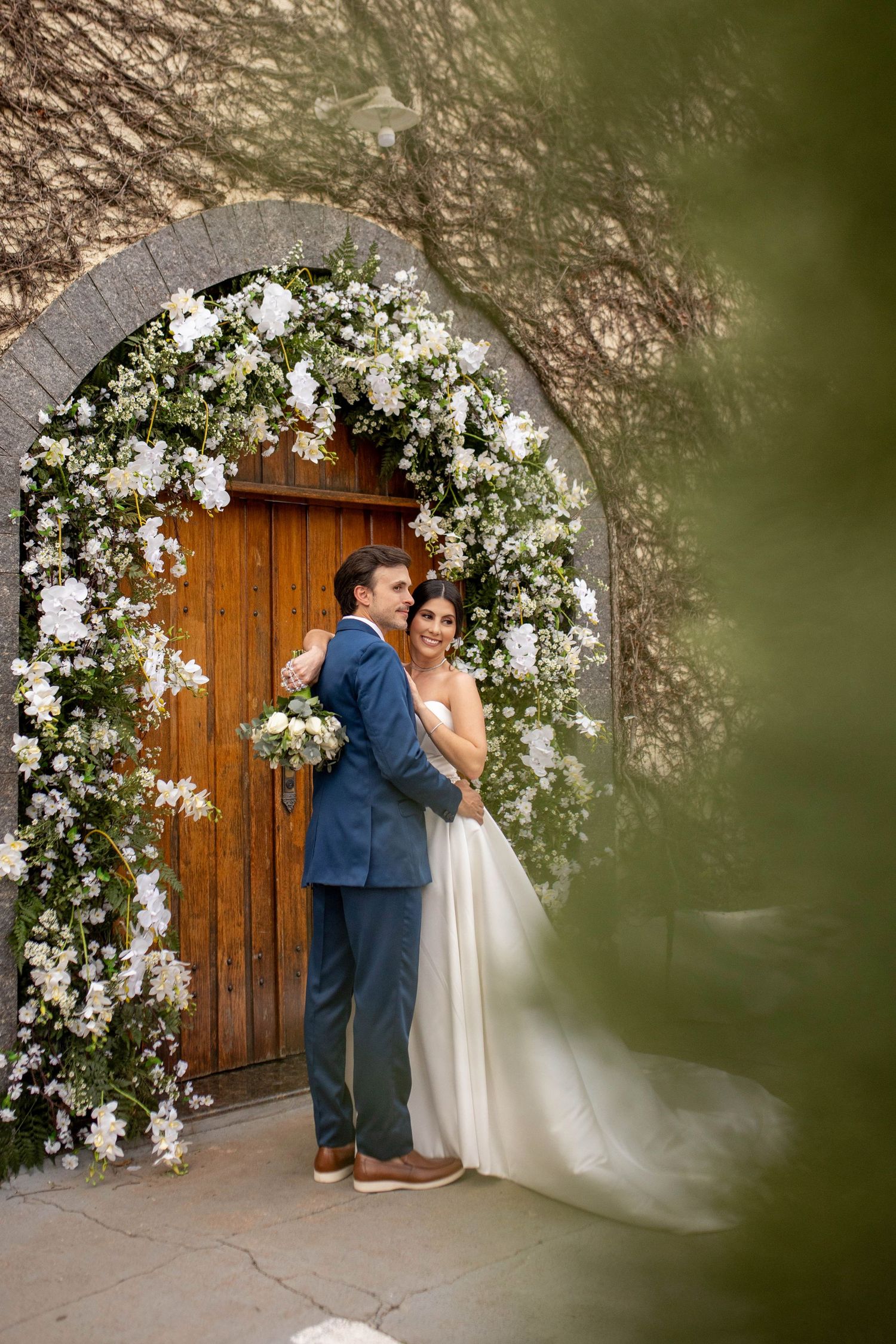 Bride and groom embrace under a floral arch with white blooms at a wooden door wedding venue.