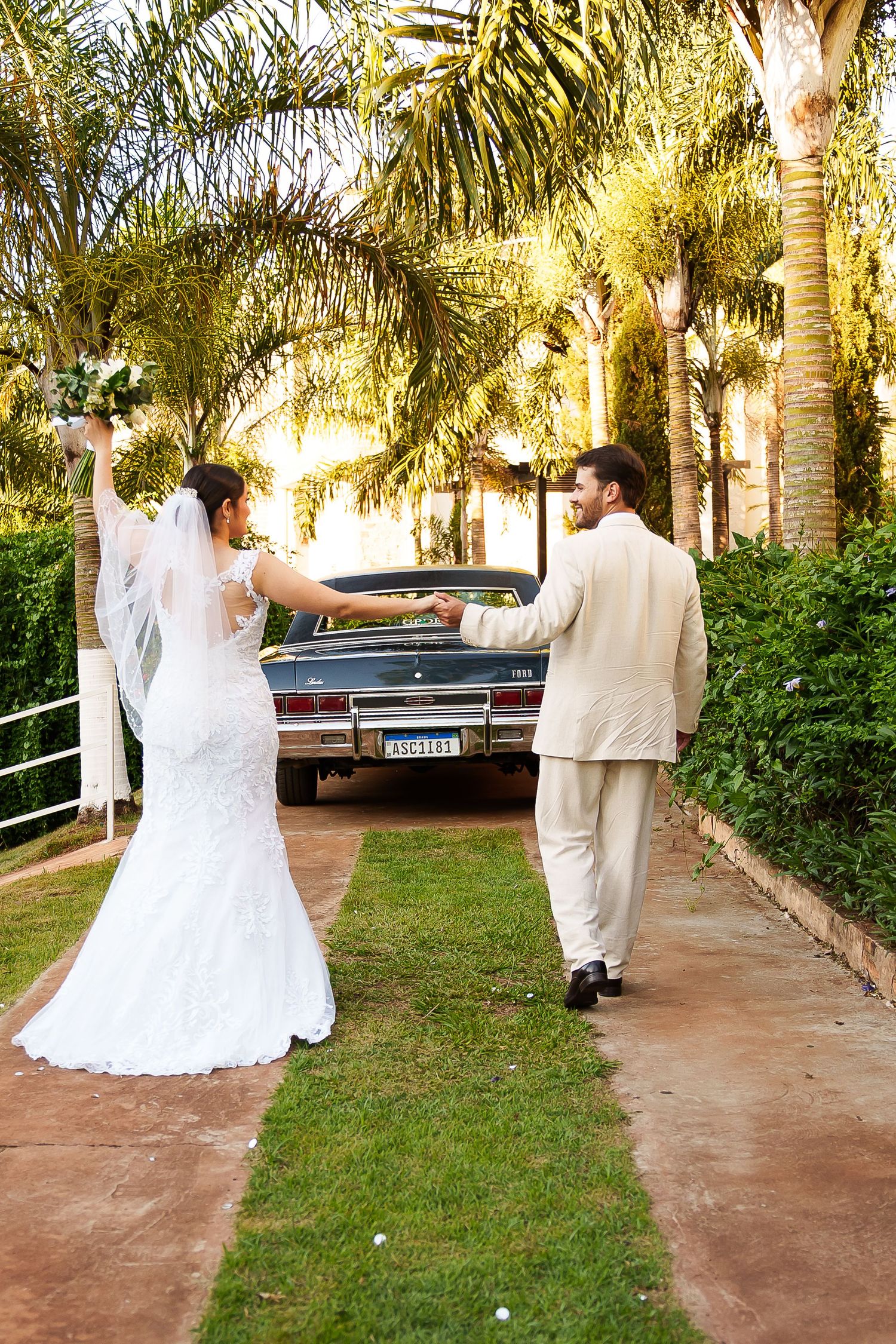 Bride and groom walking hand in hand toward a vintage car surrounded by palm trees on their wedding day.