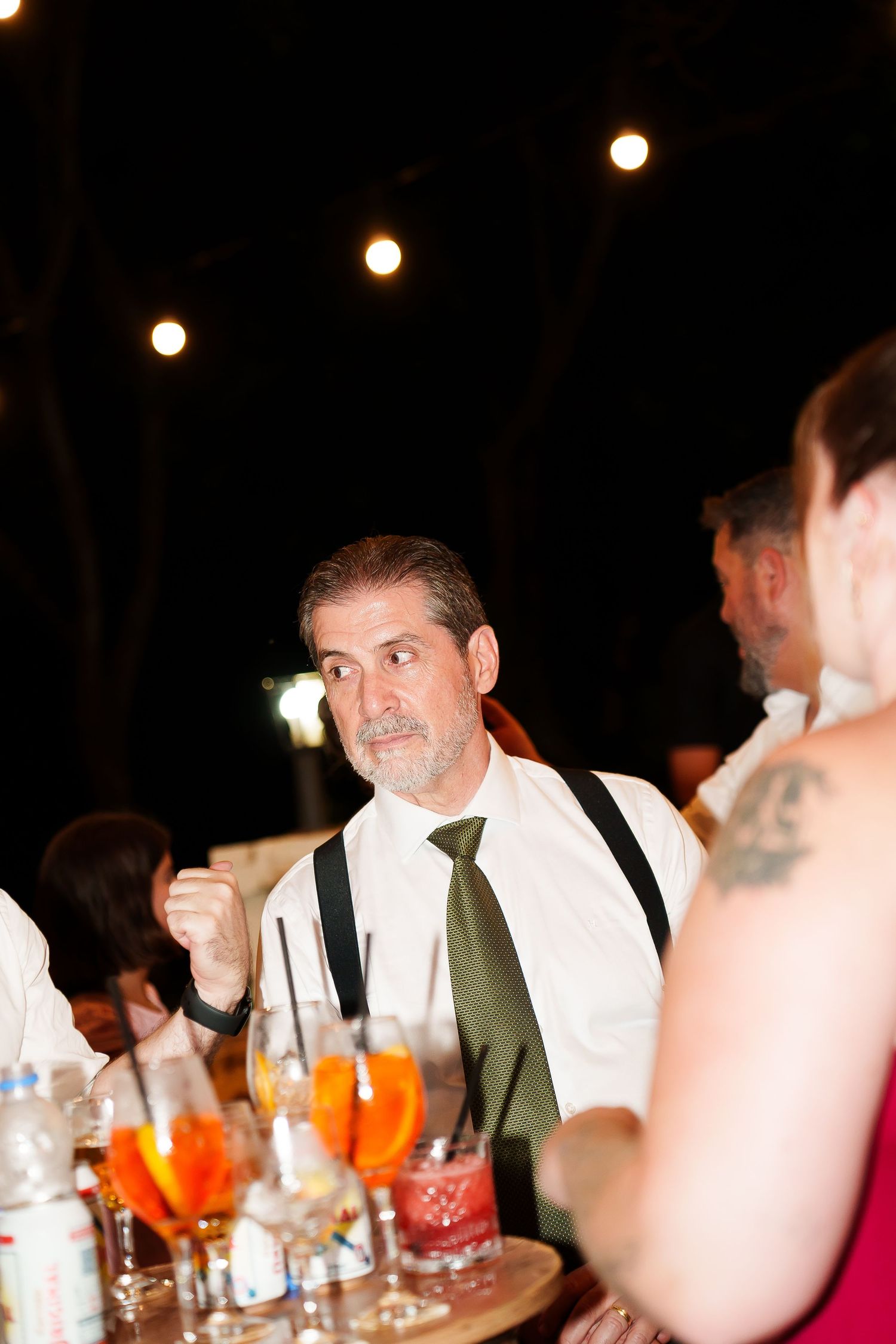 Man in white shirt, green tie, and suspenders sits at an outdoor evening event table with colorful drinks.