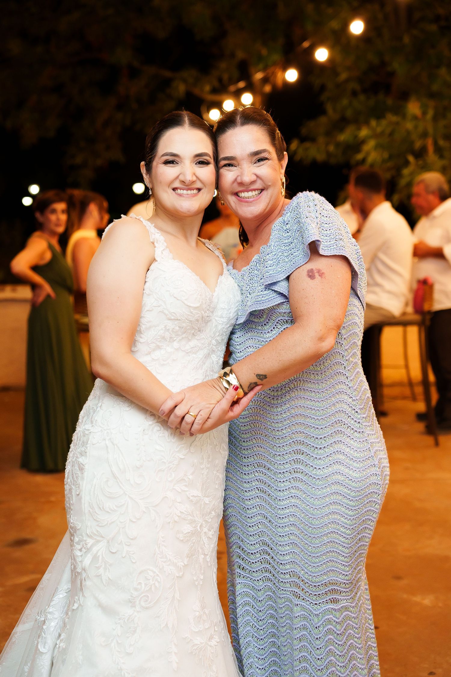 Bride in white lace gown poses with smiling woman in blue dress at outdoor wedding reception.