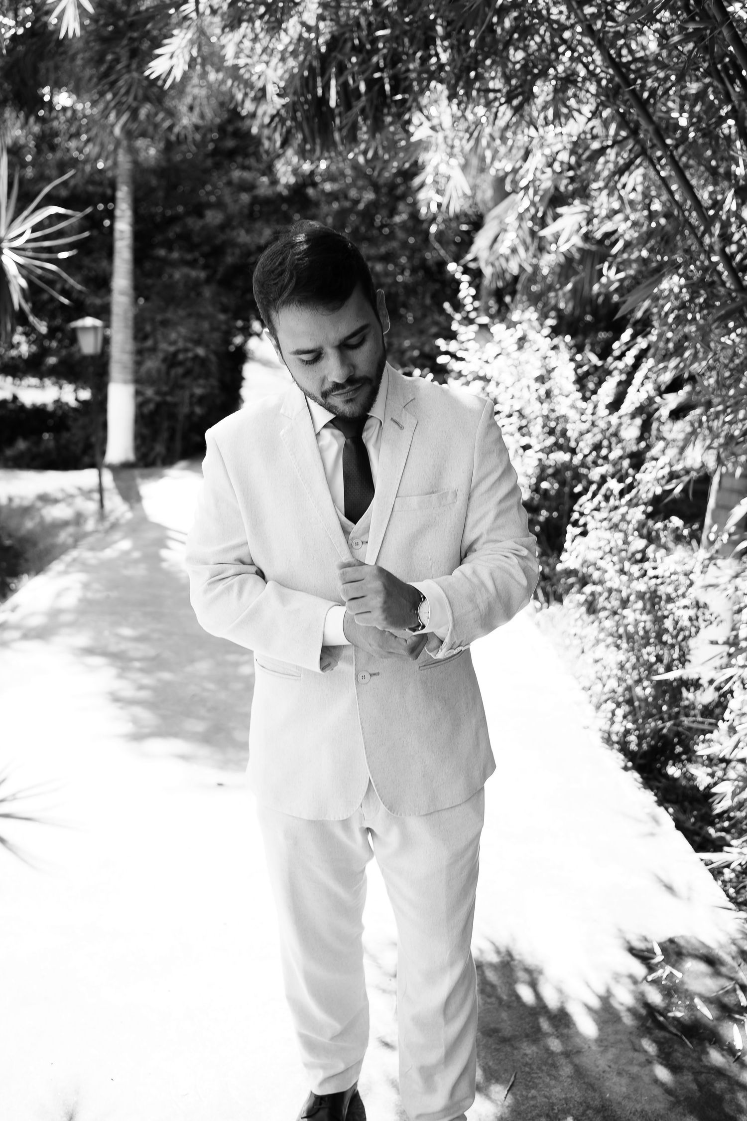 Black and white photo of a groom in a white suit adjusting his cufflinks outdoors among lush green trees.