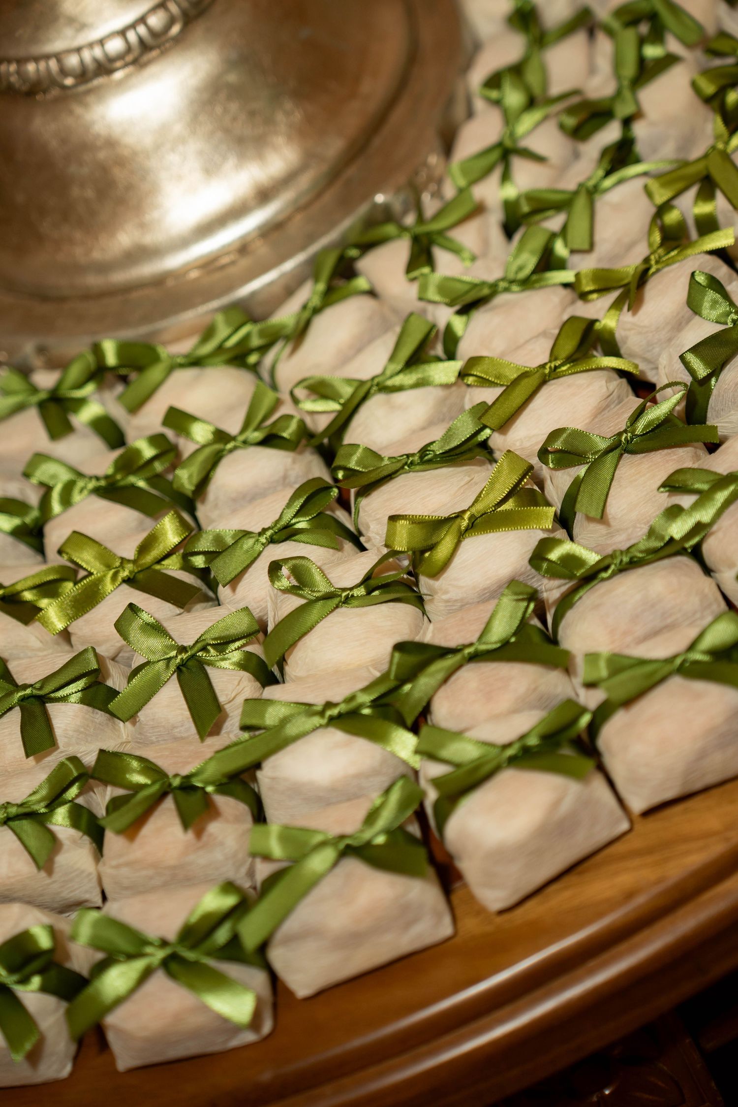 Rows of elegant gift favors wrapped in cream paper and tied with green satin ribbon bows displayed on a wooden tray.
