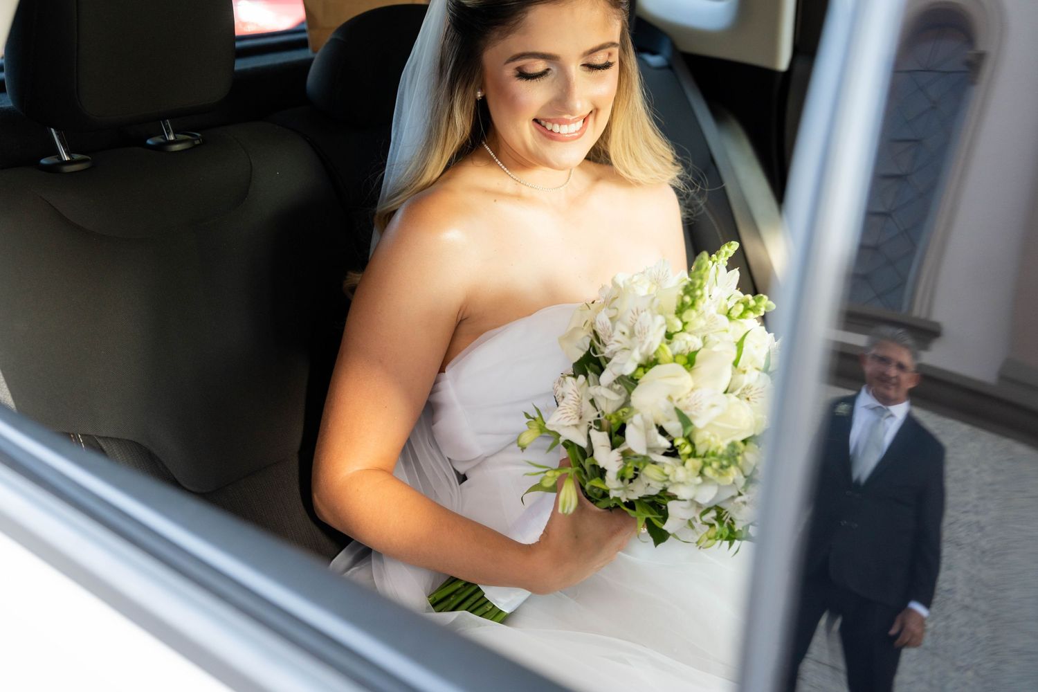 Smiling bride in white strapless gown holds white floral bouquet in car, groom visible in background reflection.