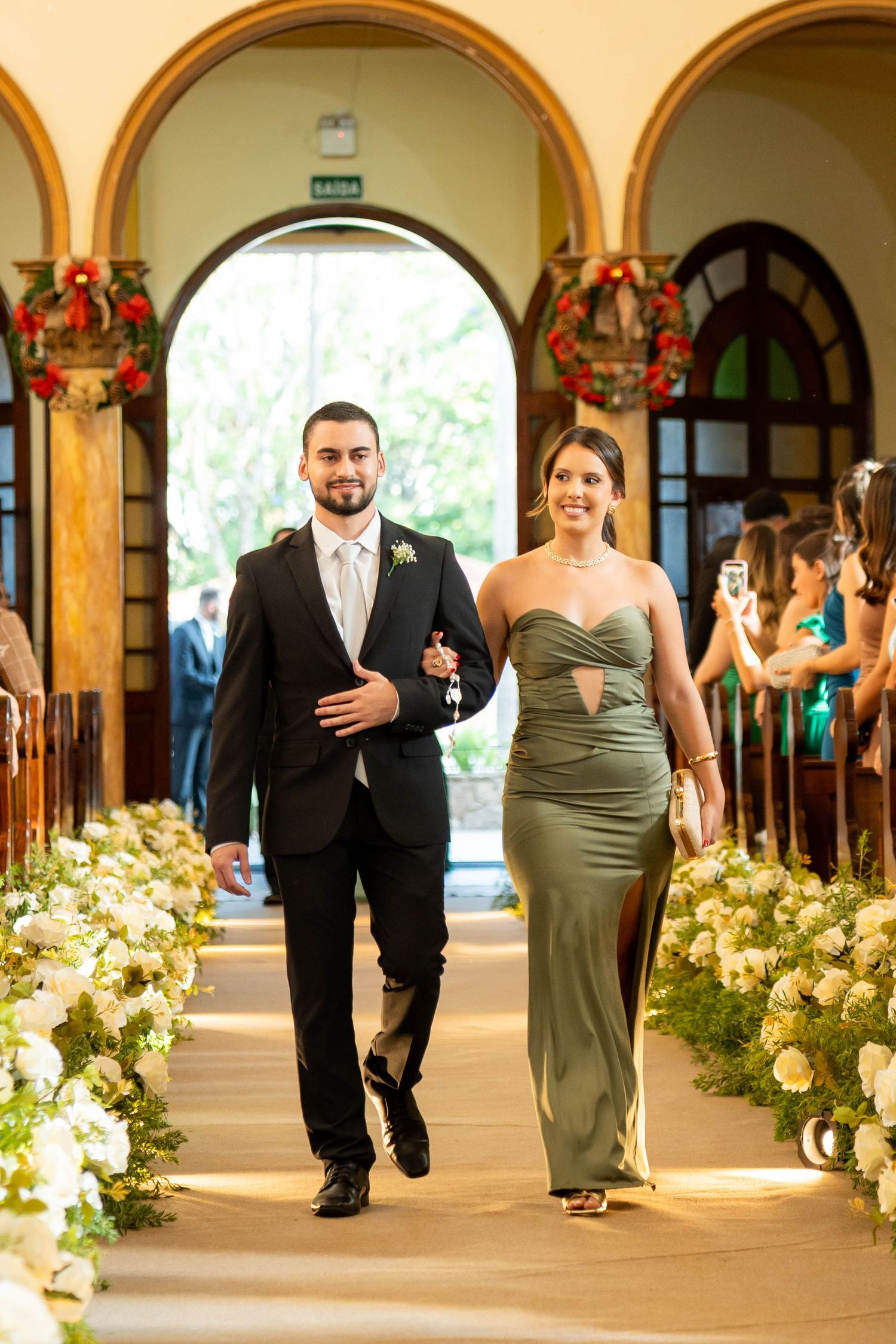 A man in a black suit and woman in an olive green gown walk down a flower-lined church aisle at a wedding.