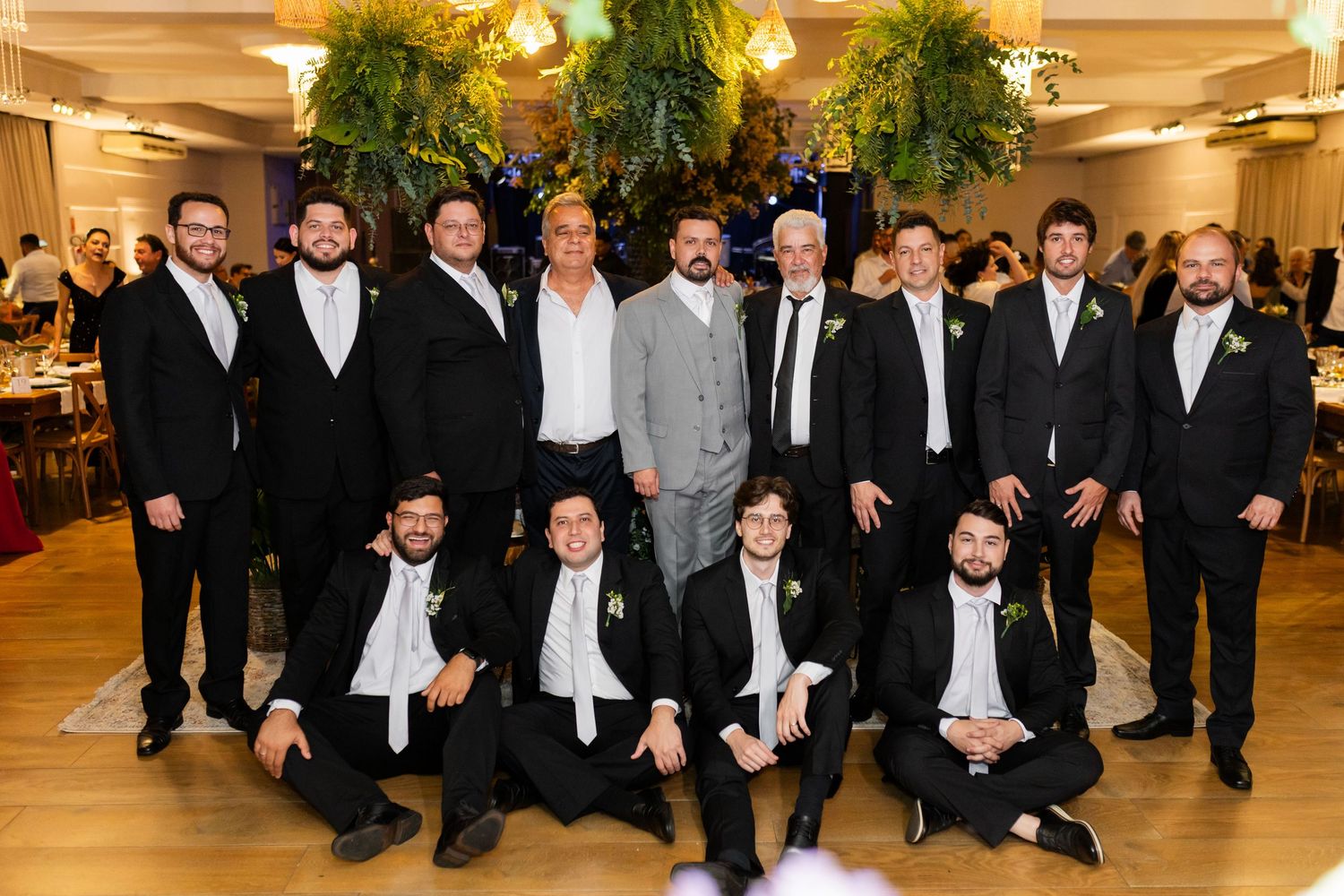 Group of men in formal black suits and one in gray suit posing together at an elegant wedding reception venue.