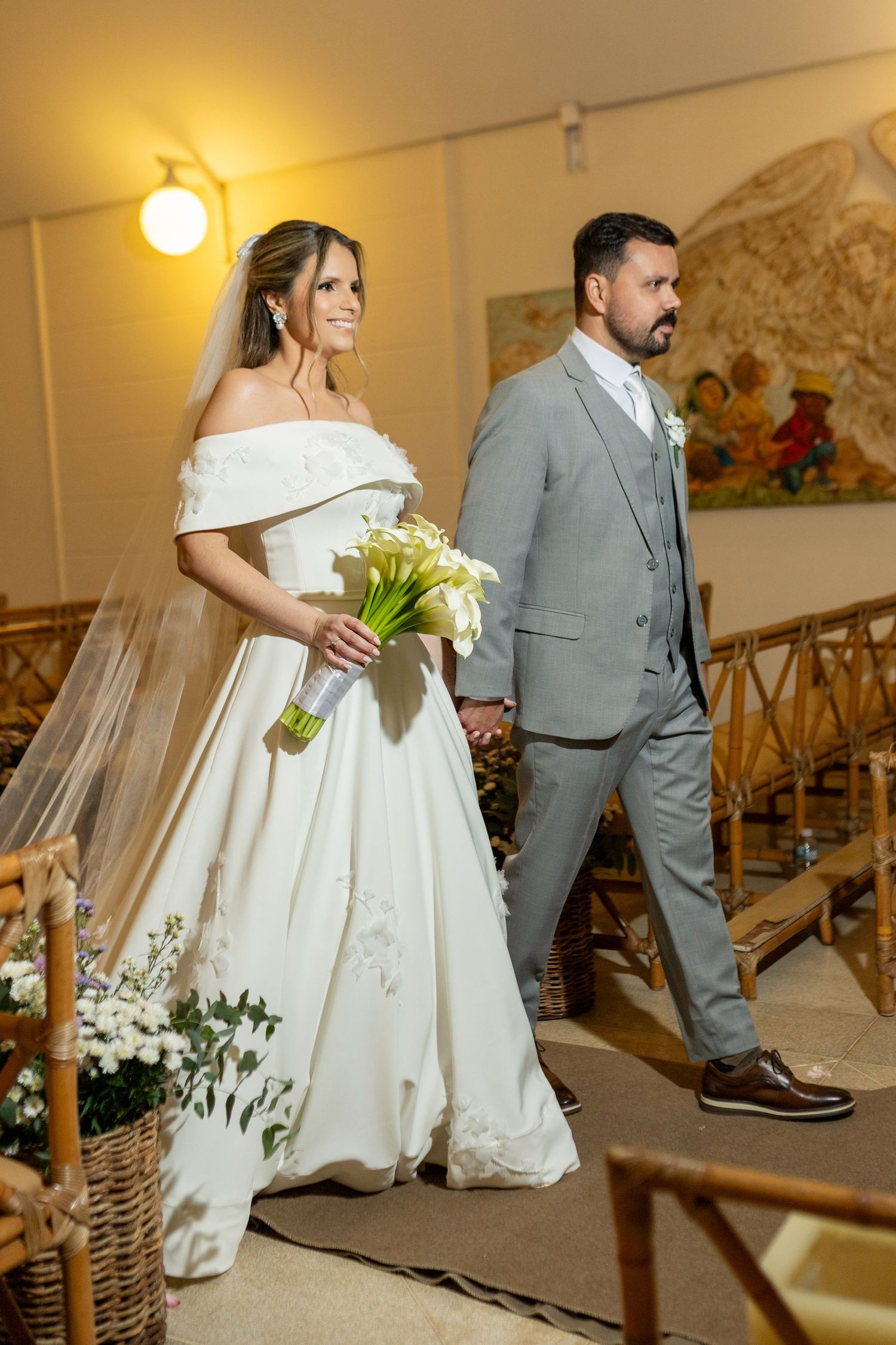 Bride in white off-shoulder gown holding calla lily bouquet walks down aisle with groom in gray suit.