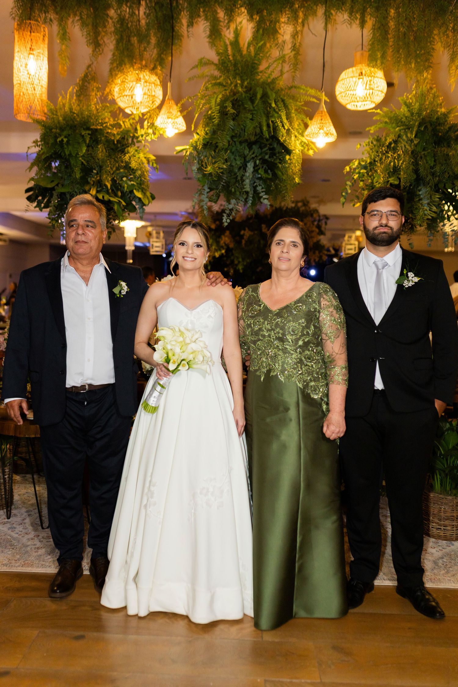Bride in white gown with bouquet poses alongside groom in tuxedo and parents at elegant green-decorated wedding venue.
