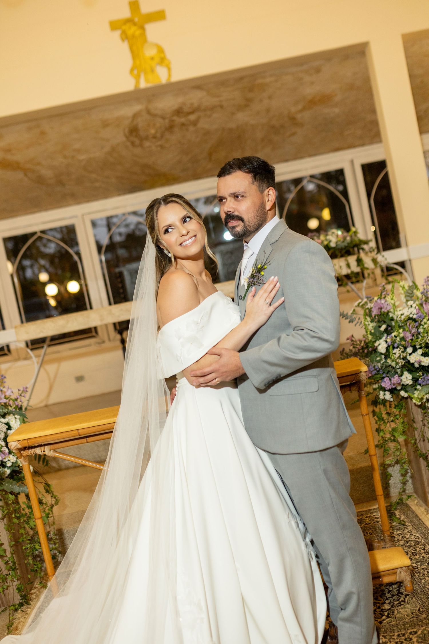 Bride and groom posing together in a church, bride in white gown with veil, groom in gray suit with boutonniere.