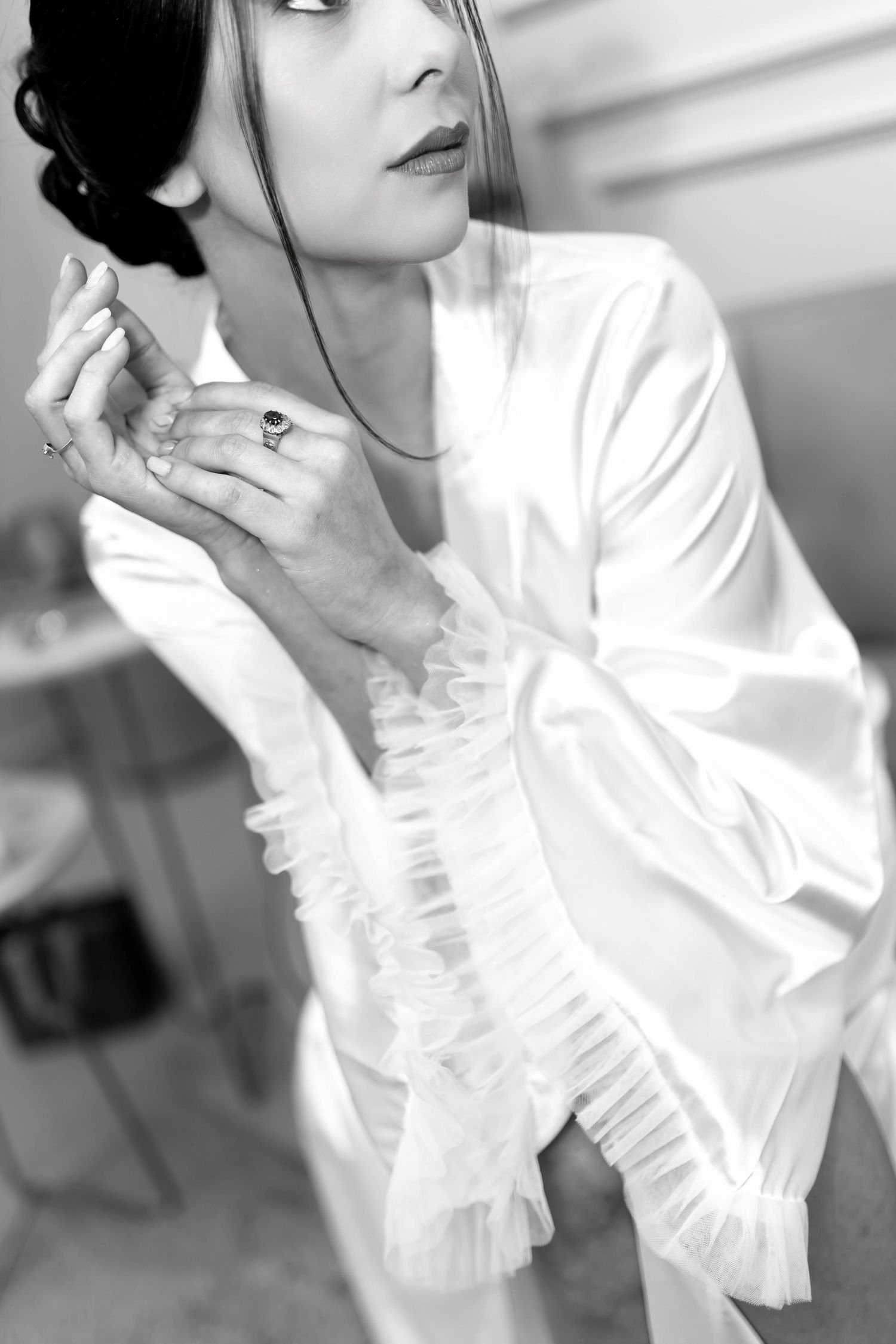 Black and white photo of elegant bride in white satin robe with ruffles, wearing rings, posing gracefully before wedding.