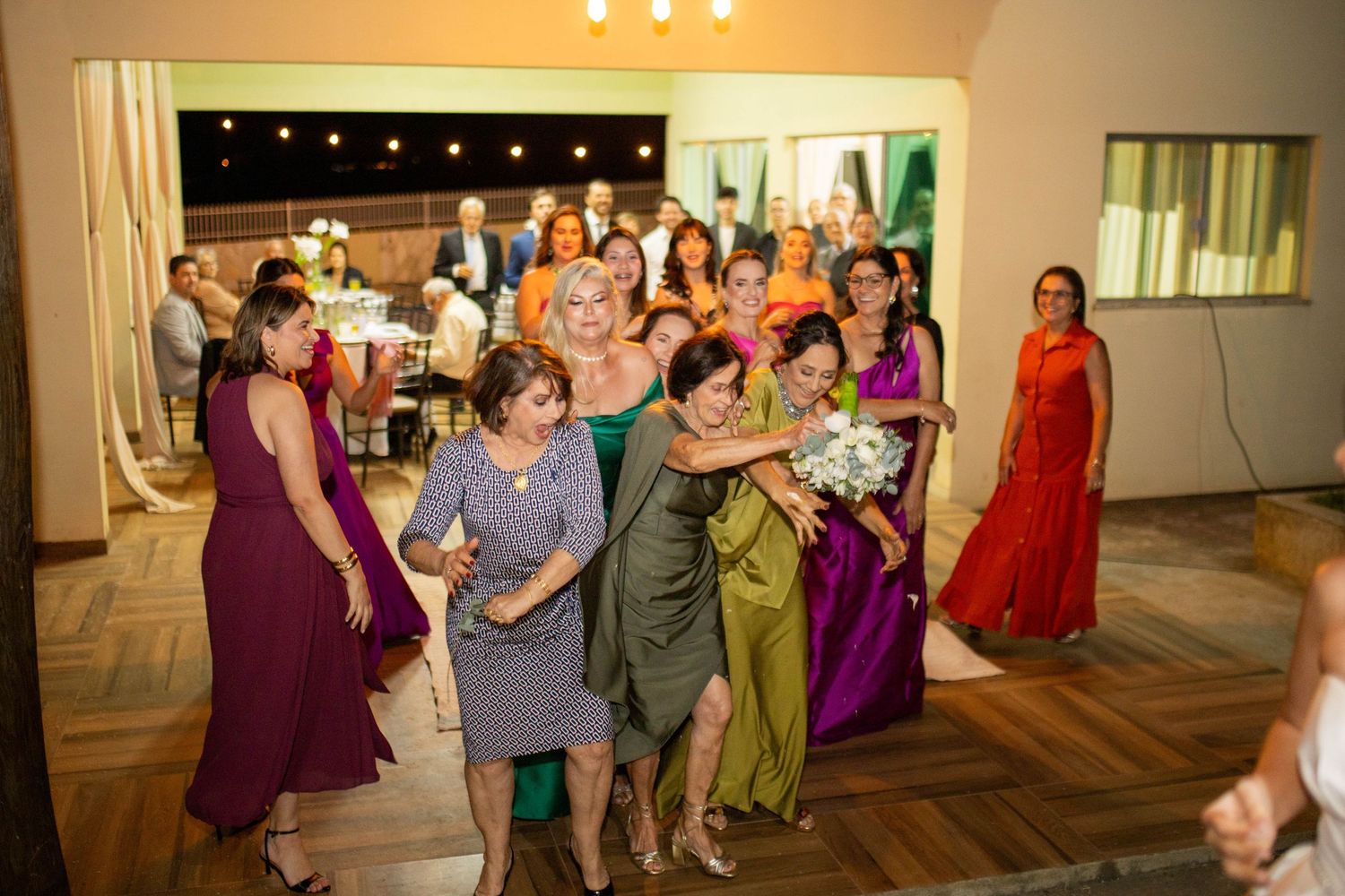 Women in colorful formal dresses gather on a dance floor during a wedding reception bouquet toss celebration.