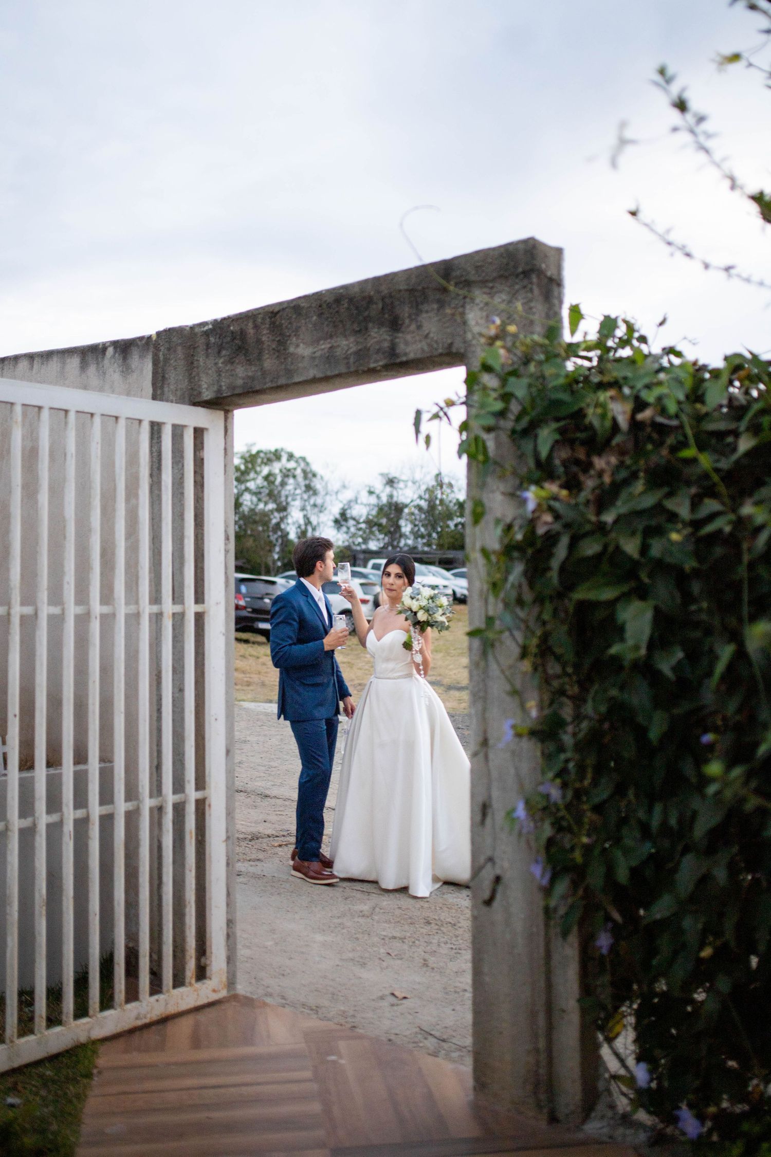 Bride in white gown and groom in blue suit stand in stone archway doorway at outdoor wedding venue.