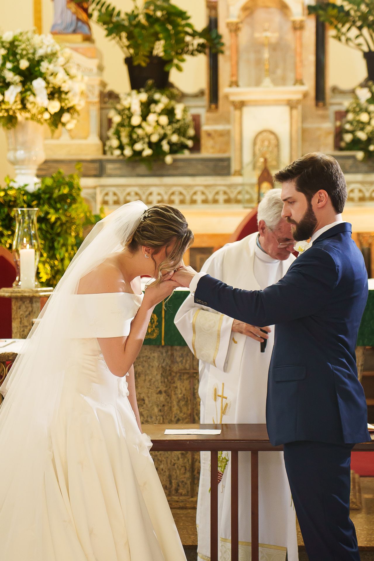 Bride kissing groom's hand during romantic church wedding ceremony with priest officiating at ornate altar.