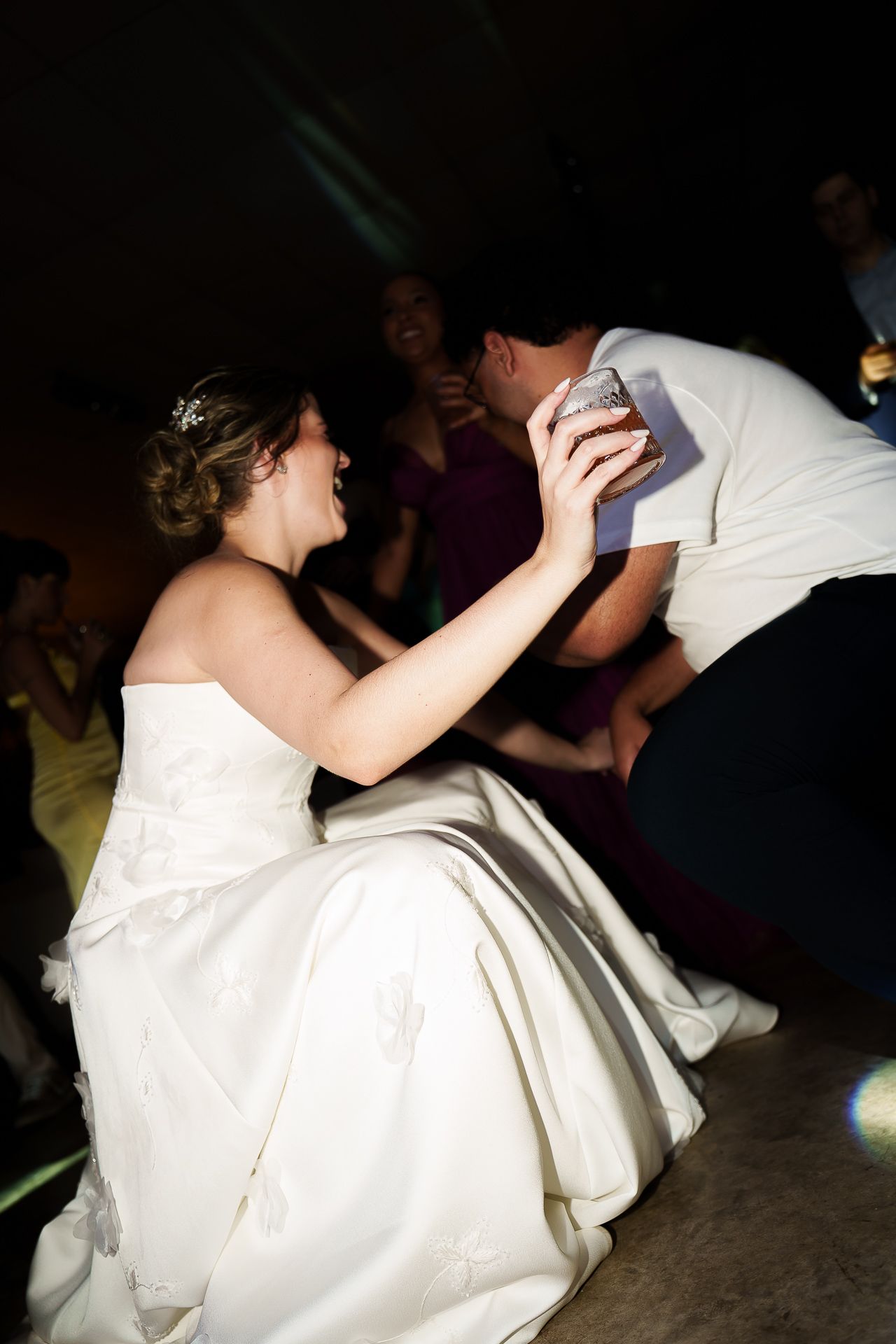 Bride in white gown laughing while groom retrieves garter during lively wedding reception dance floor celebration.