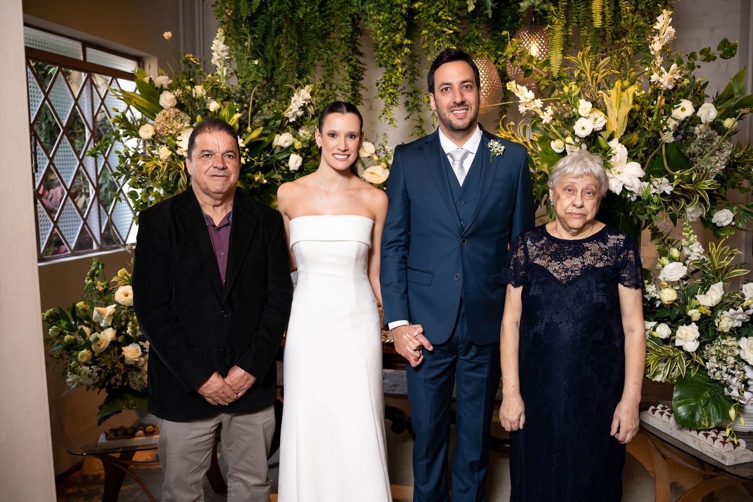Wedding party of four posing before floral backdrop, bride in white gown, groom in navy suit, flanked by older relatives.