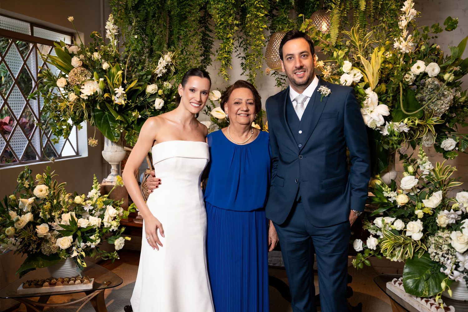 Bride in white gown, woman in blue dress, and groom in suit posing together at a floral wedding reception.