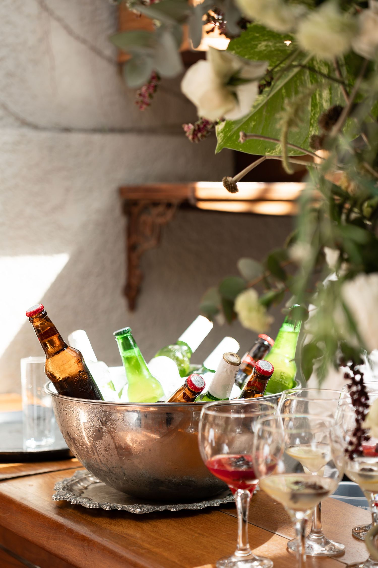 Silver ice bucket with beer bottles on wooden table, wine glasses and green foliage in elegant outdoor party setting.